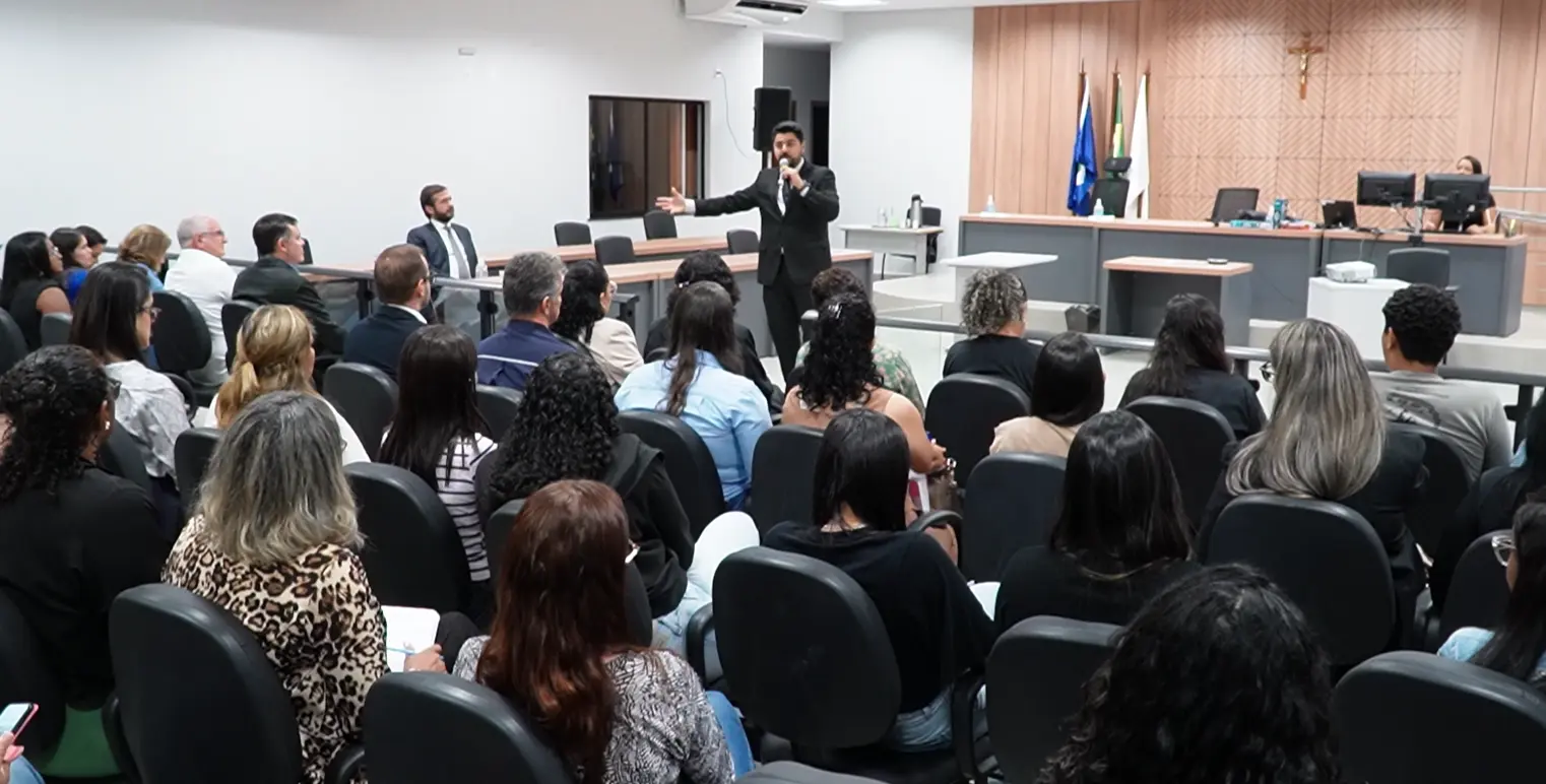 Uma sala com dezenas de pessoas sentadas assistem a um homem de terno preto que fala ao microfone, de pé, em frente a um painel de madeira com um crucifixo.