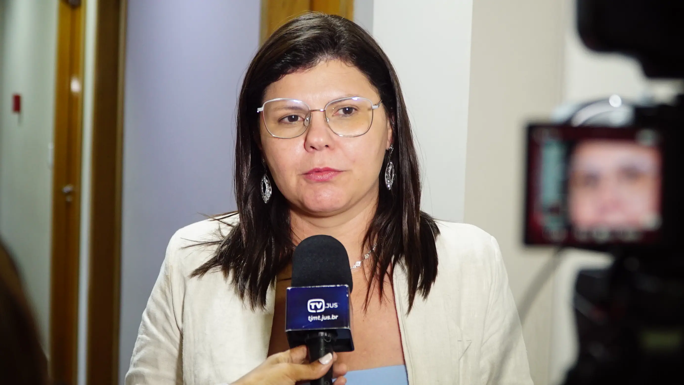 Uma mulher de cabelos castanhos lisos, óculos de armação clara e brincos longos, veste blazer bege e concede entrevista segurando microfone da TV JUS.