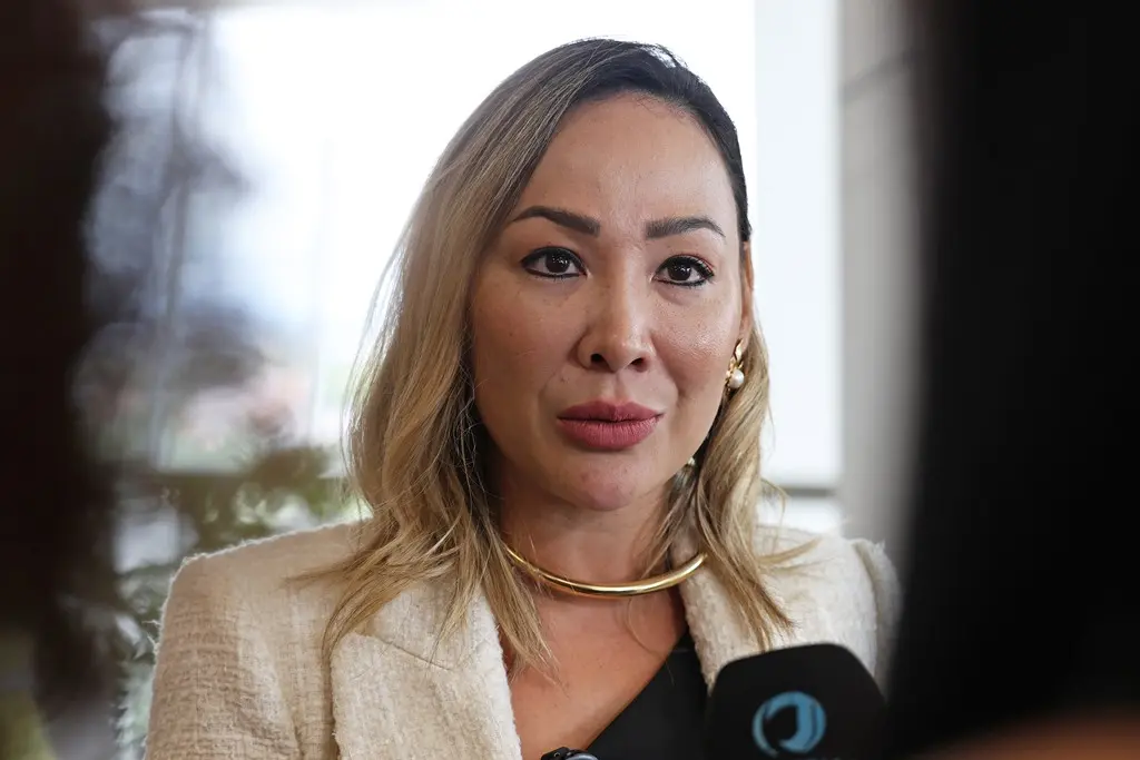 Foto horizontal em plano fechado que mostra a auditora de controle externo do Tribunal de Contas do Estado, Simony Jin, durante entrevista à TV Justiça. Ela é uma mulher de pele clara, loira, com traços orientais, usando blusa preta, blazer bege e colar de argola dourada.