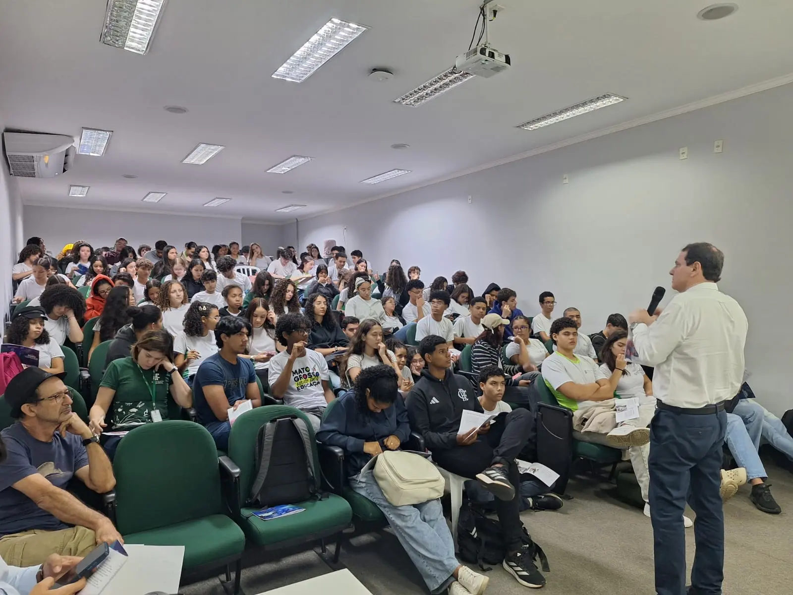 Foto horizontal que mostra dezenas de estudantes do IFMT sentados no auditório, com cartilhas do projeto Nosso Judiciário nas mãos e assistindo a uma palestra do servidor Neif Feguri, que está à frente em pé, falando ao microfone.