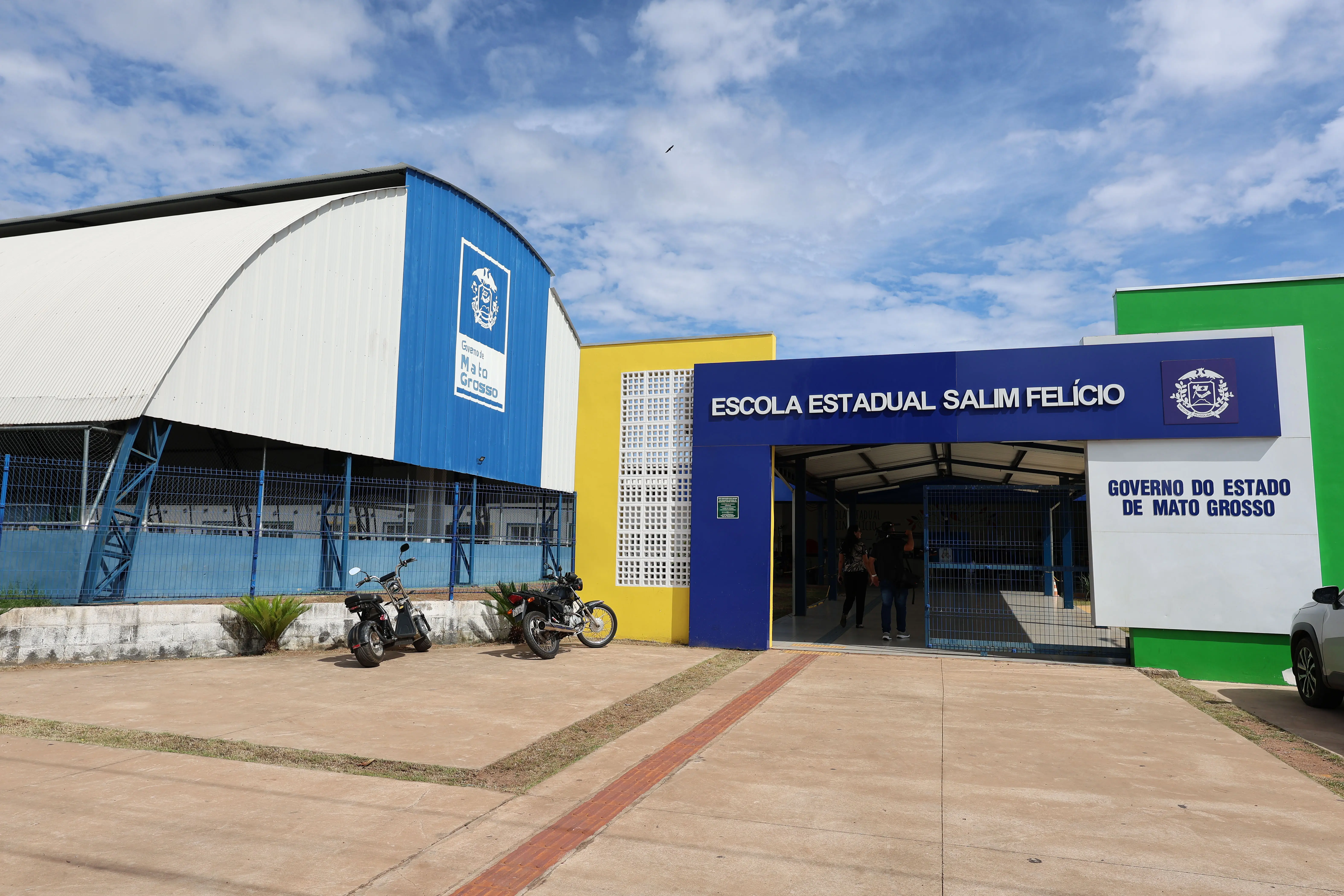 Fachada da Escola Estadual Salim Felício. Prédio moderno com as cores azul, amarelo e verde. À esquerda, um grande ginásio azul com o brasão de Mato Grosso. Na entrada, letreiro branco sobre fundo azul e motocicletas estacionadas.