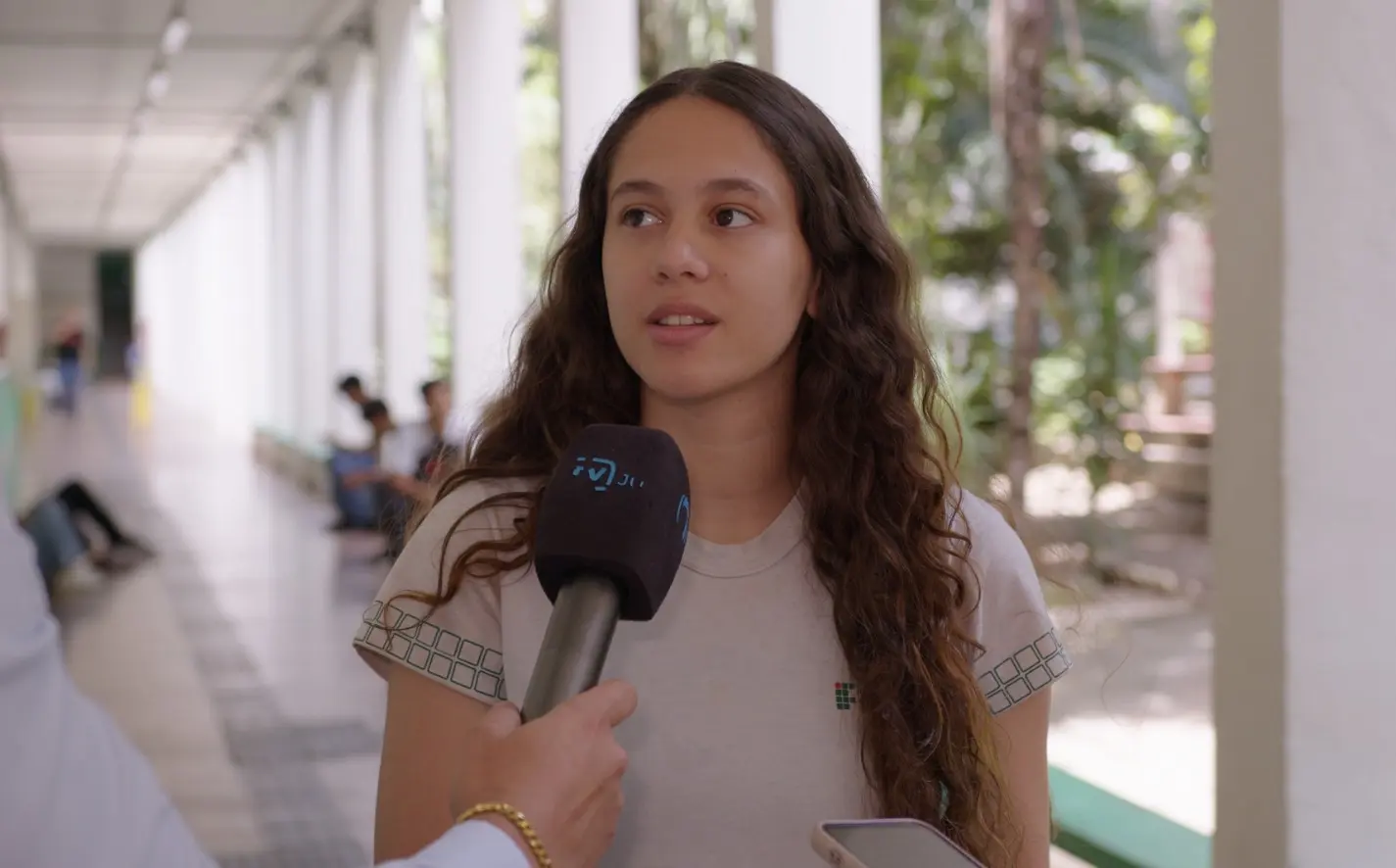 Foto horizontal em plano médio que mostra a estudante Yasmim Vitória durante entrevista à TV Justiça, no corredor do IFMT. Ela é uma adolescente de pele branca, magra, cabelos cacheados longos e soltos, usando camiseta branca da escola.