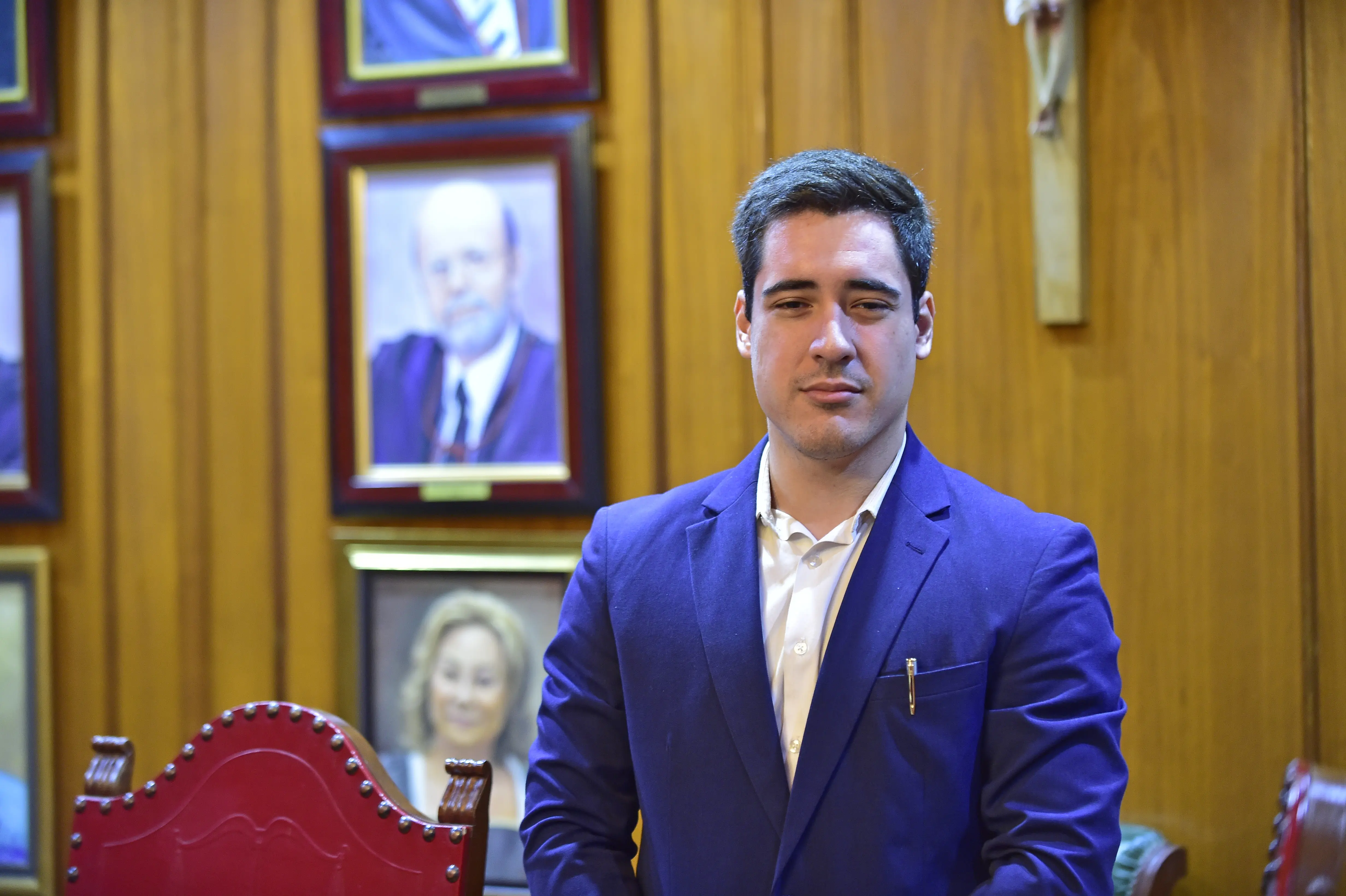 o Aluno Daniel Bottassini está em destaque na imagem. Ele é um homem branco de cabelos negros, que veste um terno azul-marinho sobre uma camisa branca. Ele está em pé. Atrás dele há quadros com imagens de desembargadores do TJMT e uma parte de um crucifixo. 