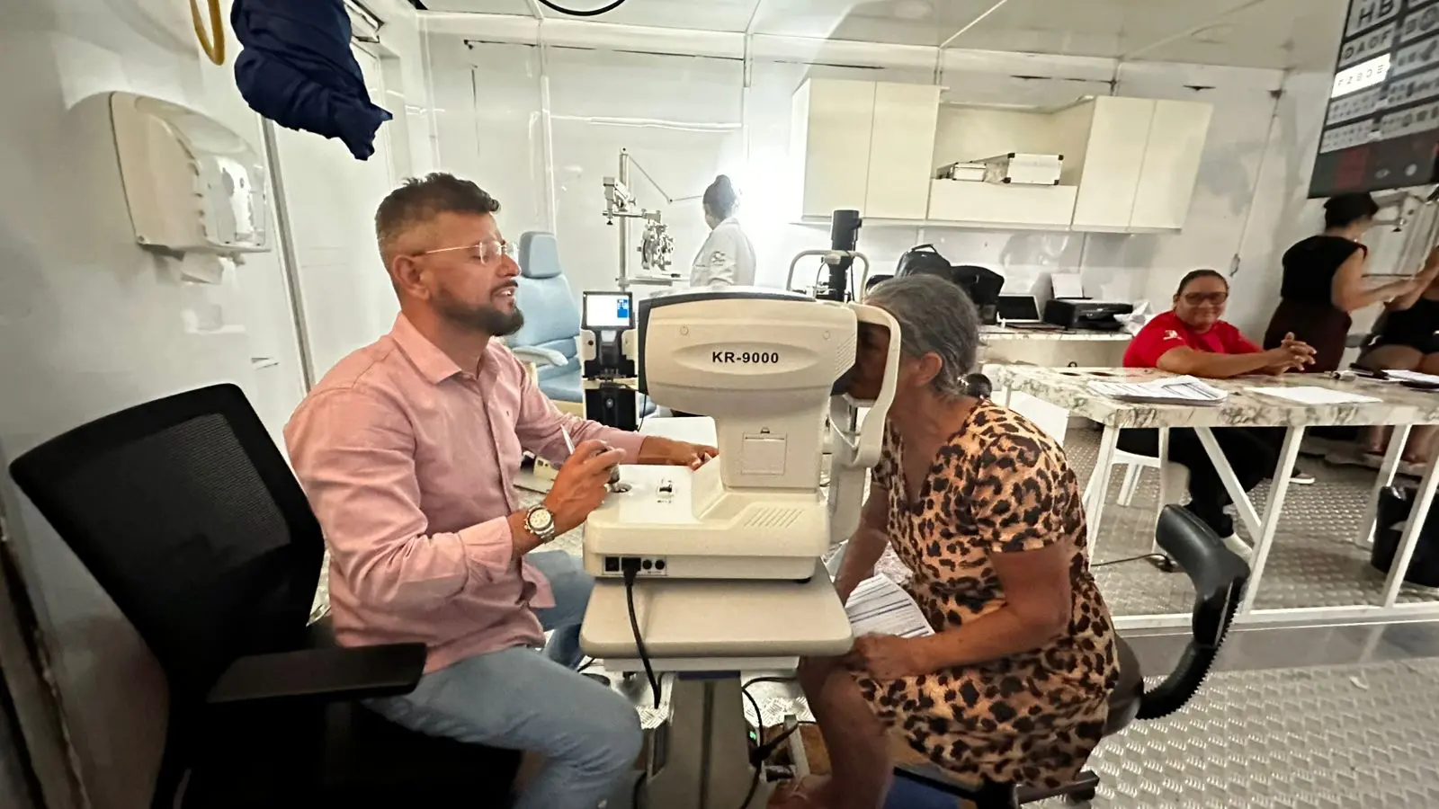 Profissional de saúde masculino realiza exame oftalmológico em senhora idosa utilizando equipamento digital. Ao fundo, outros atendimentos acontecem simultaneamente, demonstrando a estrutura multisserviços.