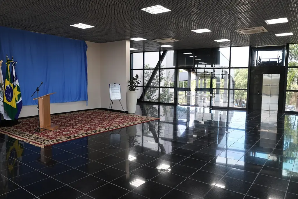 Foto na horizontal do hall de entrada do Plenário 1 do Tribunal de Justiça de Mato Grosso. O espaço é amplo, com piso preto, janelas de vidro, um grande tapete e acesso por elevador e rampa.
