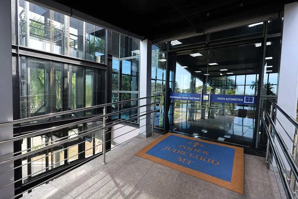 Foto na horizontal que mostra uma a entrada do Plenário do Tribunal de Justiça pela rampa de acesso. A porta e de vidro e tem um grande tapete azul e amarelo escrito Poder Judiciário MT. A rampa tem corrimões metálicos.