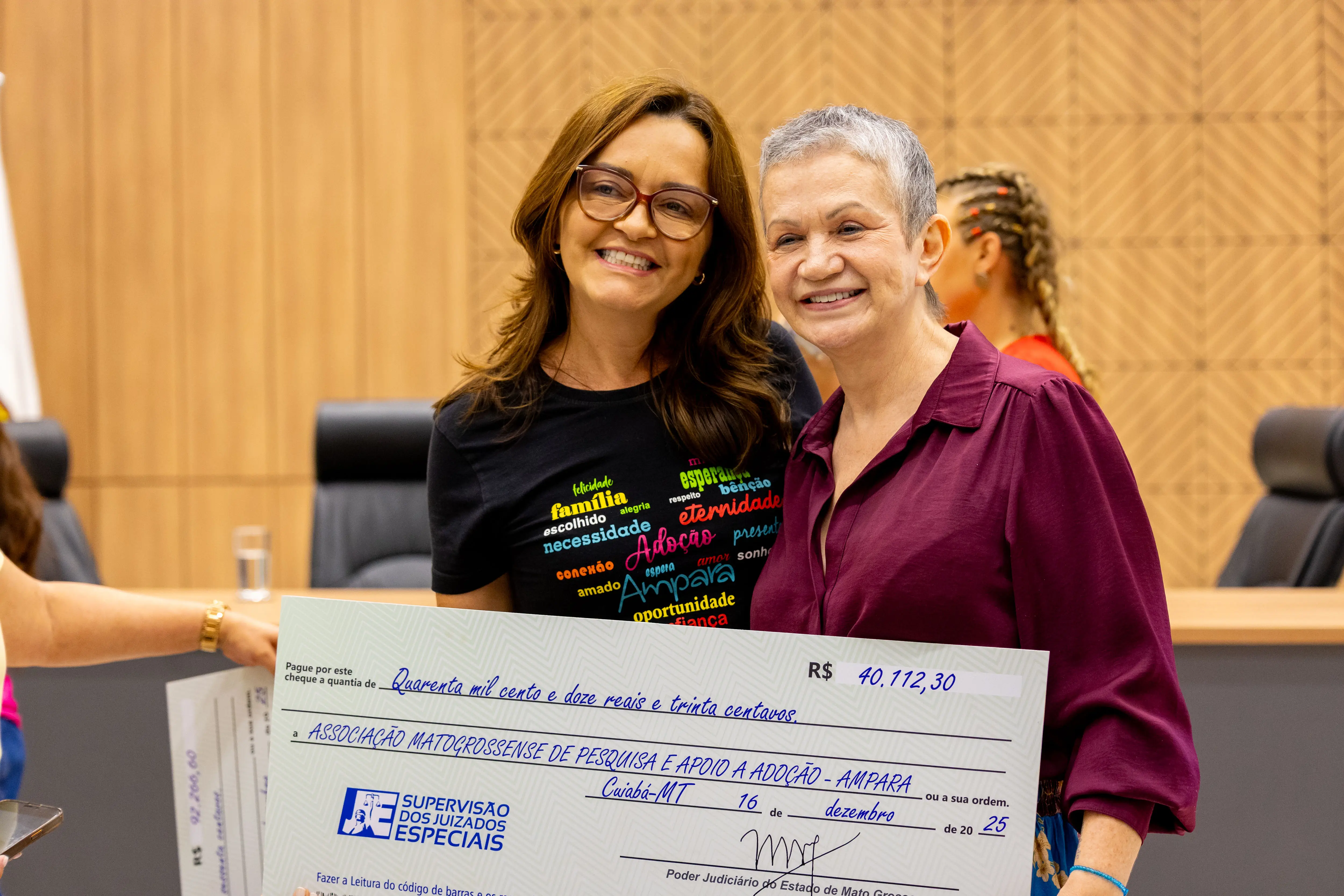Duas mulheres sorriem segurando cheque simbólico em ambiente institucional. Uma usa camiseta preta com palavras coloridas, a outra veste blusa vinho e tem cabelos curtos grisalhos. Ao fundo, mesa oficial e painel de madeira.