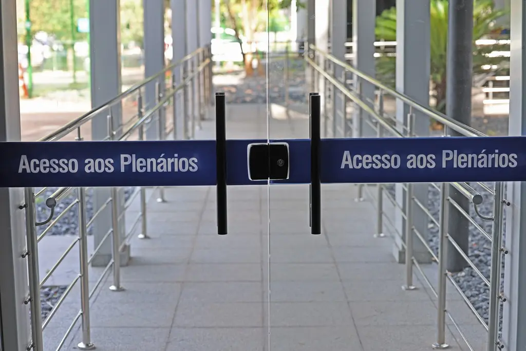 foto horizontal que mostra uma porta de vidro com uma fita adesiva azul onde está escrito "Acesso aos Plenários". do outro lado do vidro, é possível ver um caminho de piso de concreto e corrimões metálicos.