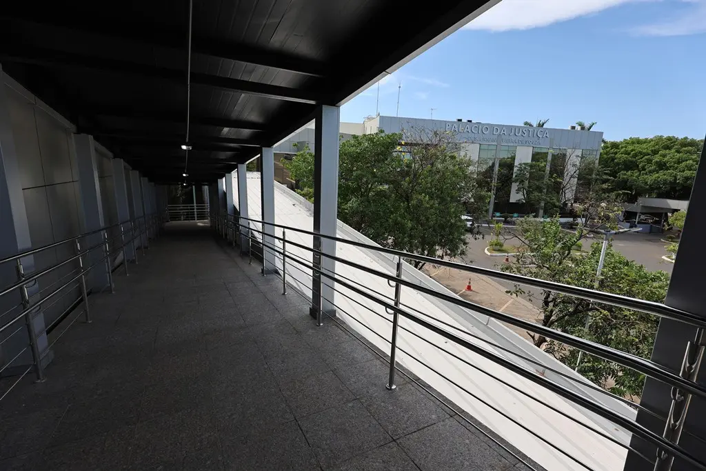 Foto na horizontal que mostra a rampa de acesso ao Plenário 1 do Tribunal de Justiça de Mato Grosso. A rampa é de concreto, coberta e com corrimões metálicos. Na lateral direita, é possível ver a fachada do prédio e várias árvores.