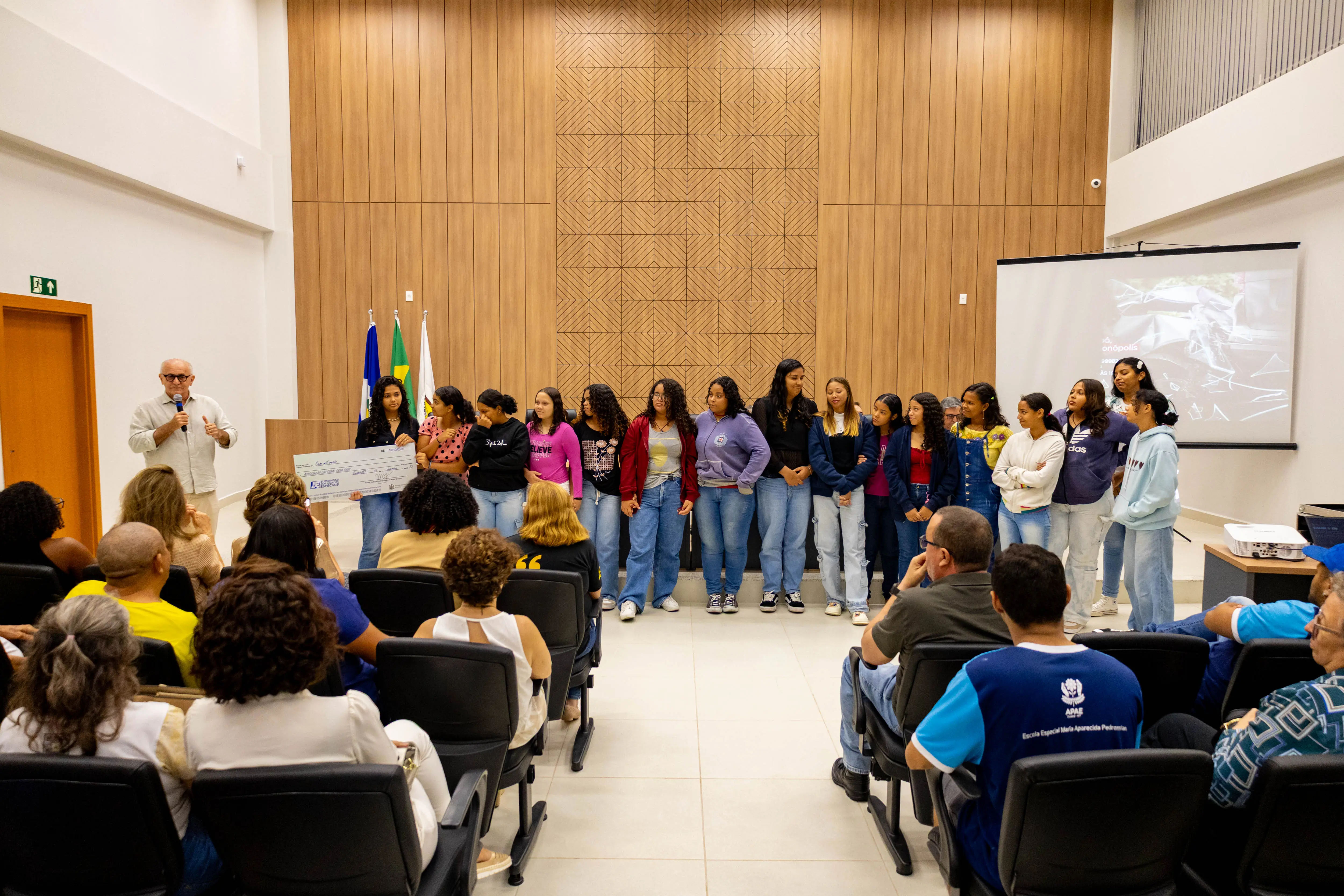 Vista geral do auditório com público sentado e grupo em pé à frente. Um homem fala ao microfone enquanto jovens alinham-se ao lado. Telão e bandeiras reforçam o caráter institucional do evento.