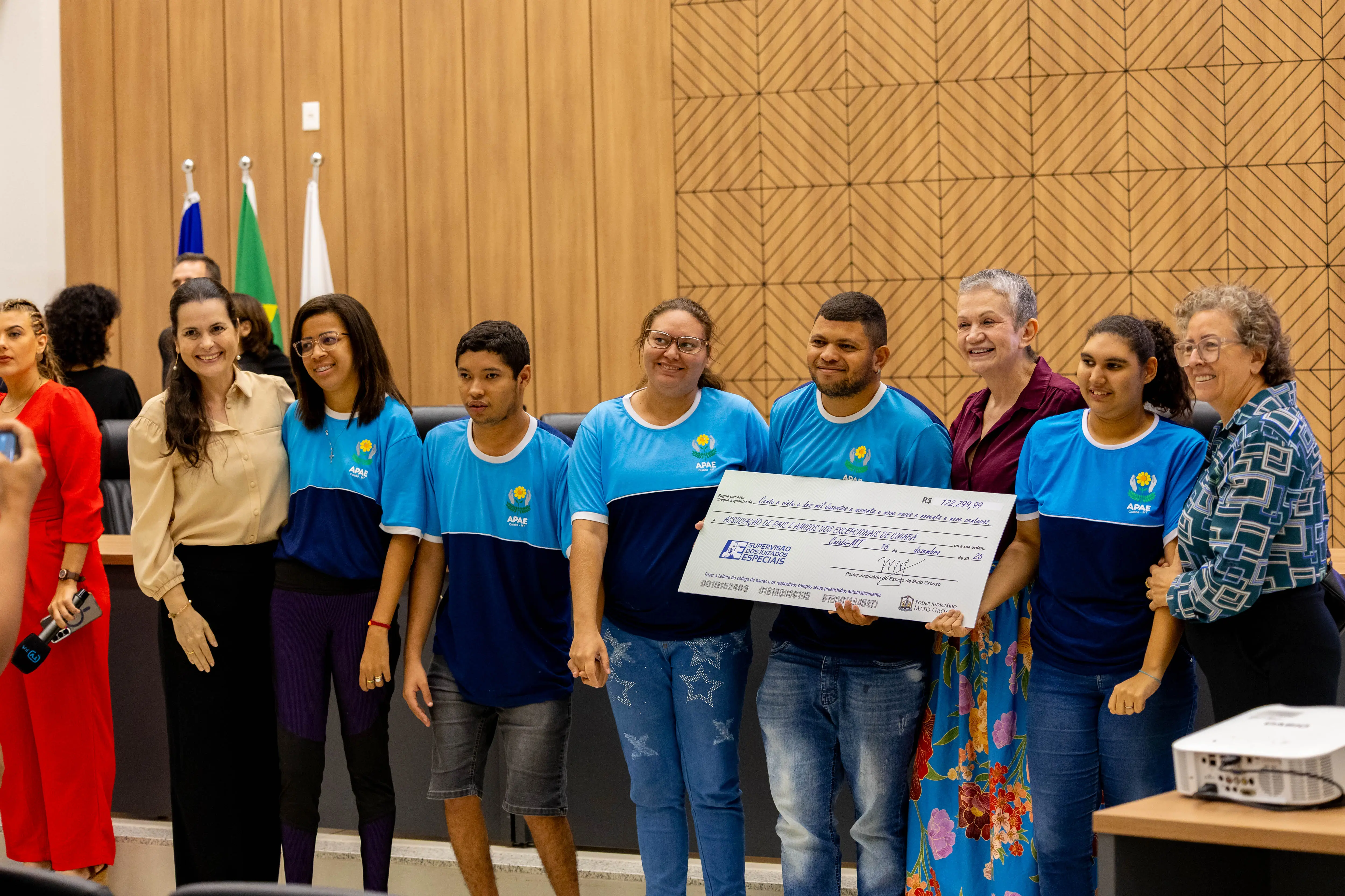 Grupo de homens e mulheres, a maioria com camisetas azuis da Apae, posa em auditório segurando cheque simbólico. Todos sorriem, em frente a mesa institucional, com bandeiras e painel de madeira ao fundo.