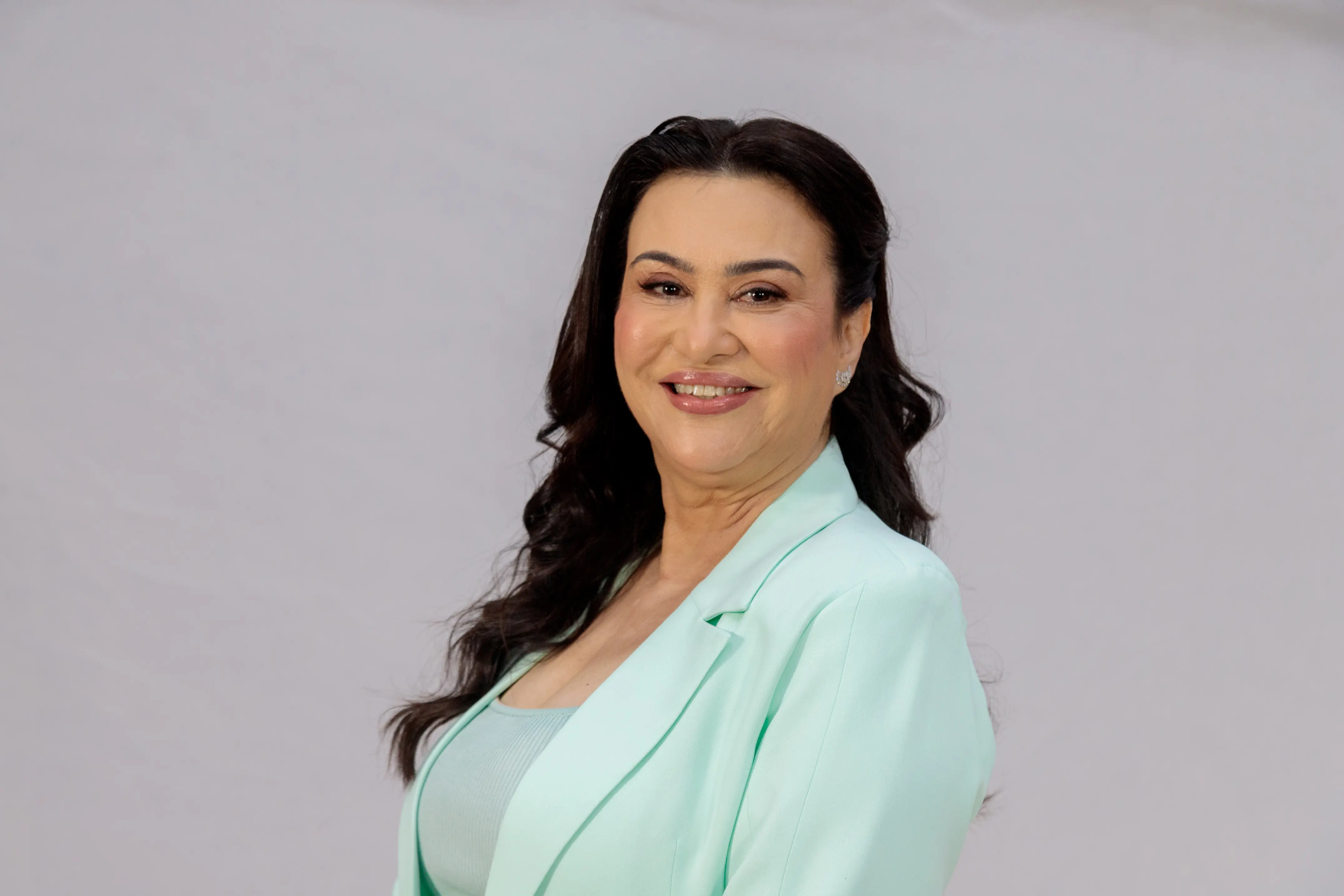 Desembargadora Anglizey de Oliveira. Ela posa sorridente em um retrato oficial, exibindo longos cabelos escuros ondulados e maquiagem suave. Ela veste um blazer em tom verde-água sobre uma blusa de cor semelhante, transmitindo uma imagem de elegância e serenidade em um fundo neutro e claro. A compos