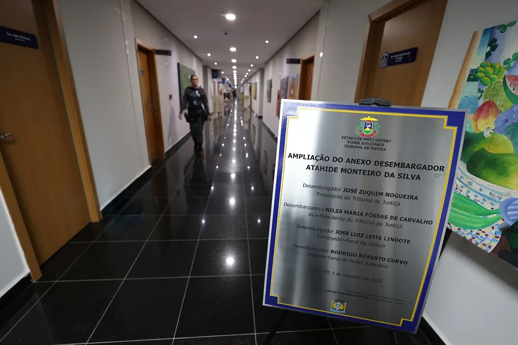 Foto horizontal de um corredor comprido, localizado dentro do Tribunal de Justiça de Mato Grosso. Esse corredor é cheio de portas de madeira, que dão acesso aos gabinetes de desembargadores. Em primeiro plano, há uma placa de inauguração de novos gabinetes e uma policial fardada anda no corredor.