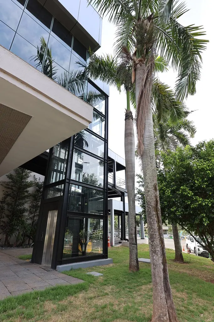 Foto vertical que mostra o exterior do elevador panorâmico que dá acesso ao Plenário 1 do Tribunal de Justiça de Mato Grosso. É um elevador de esquadrias metálicas e vidro, em um terreno gramado e com palmeiras e outras árvores em volta.
