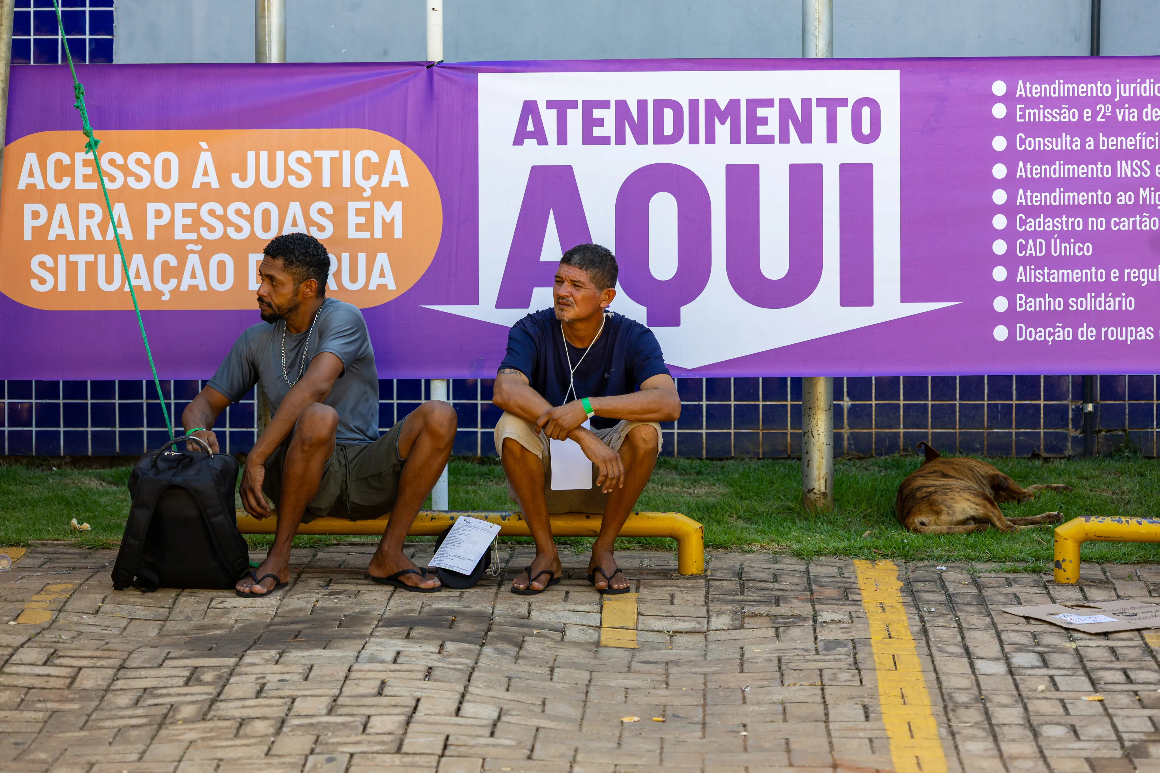Dois homens sentados em uma mureta baixa diante de banner roxo com a frase "Atendimento Aqui" e "Acesso à Justiça para Pessoas em Situação de Rua". Um cachorro dorme na grama ao lado.