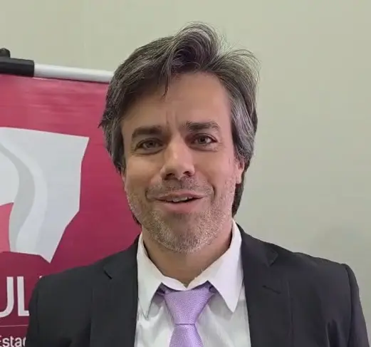 Homem branco de cabelo grisalho, com terno preto e gravata roxa, sorrindo e falando. 