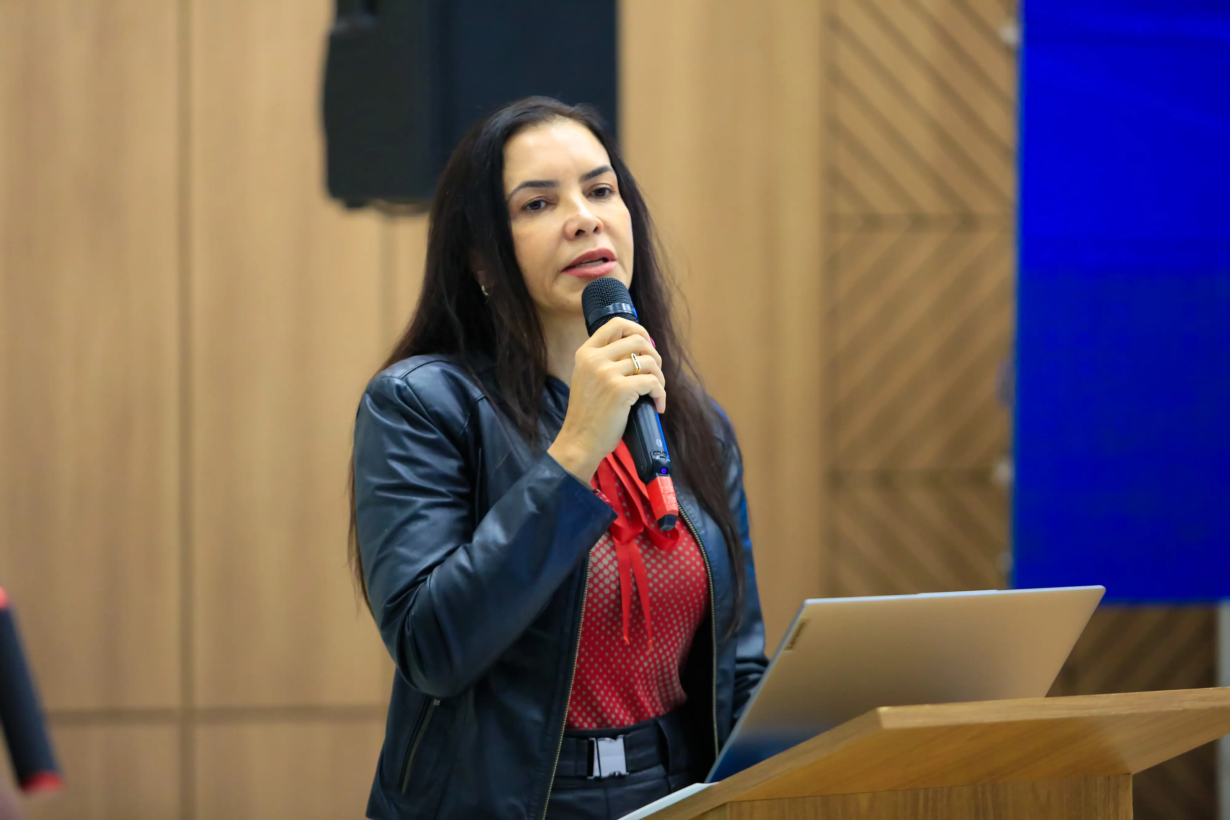 Mulher branca de cabelos longos e escuros fala ao microfone. Ela usa jaqueta de couro preta sobre blusa vermelha com poá. Está posicionada atrás de um púlpito de madeira clara, com expressão decidida durante sua fala no evento.