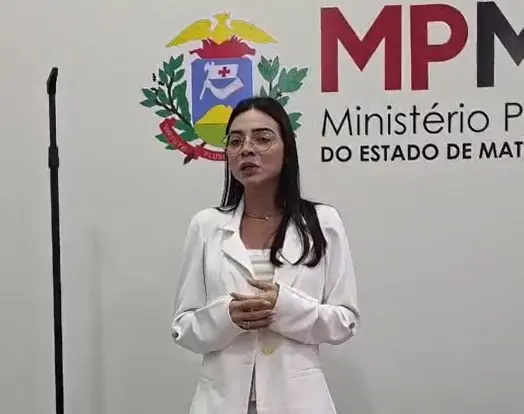 Mulher branca, de cabelos escuros longos, com óculos e blazer branco, falando em frente ao logo do MPMT na parede. 
