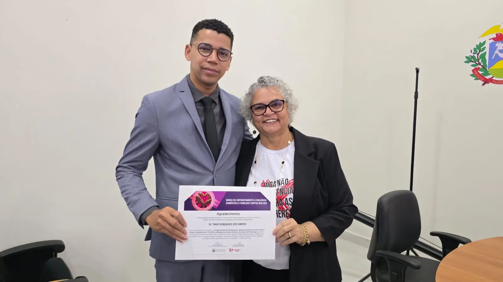 Jovem de terno cinza entregando segurando um certificado com uma mulher de cabelos grisalhos de blazer preto, com camisa branca. 