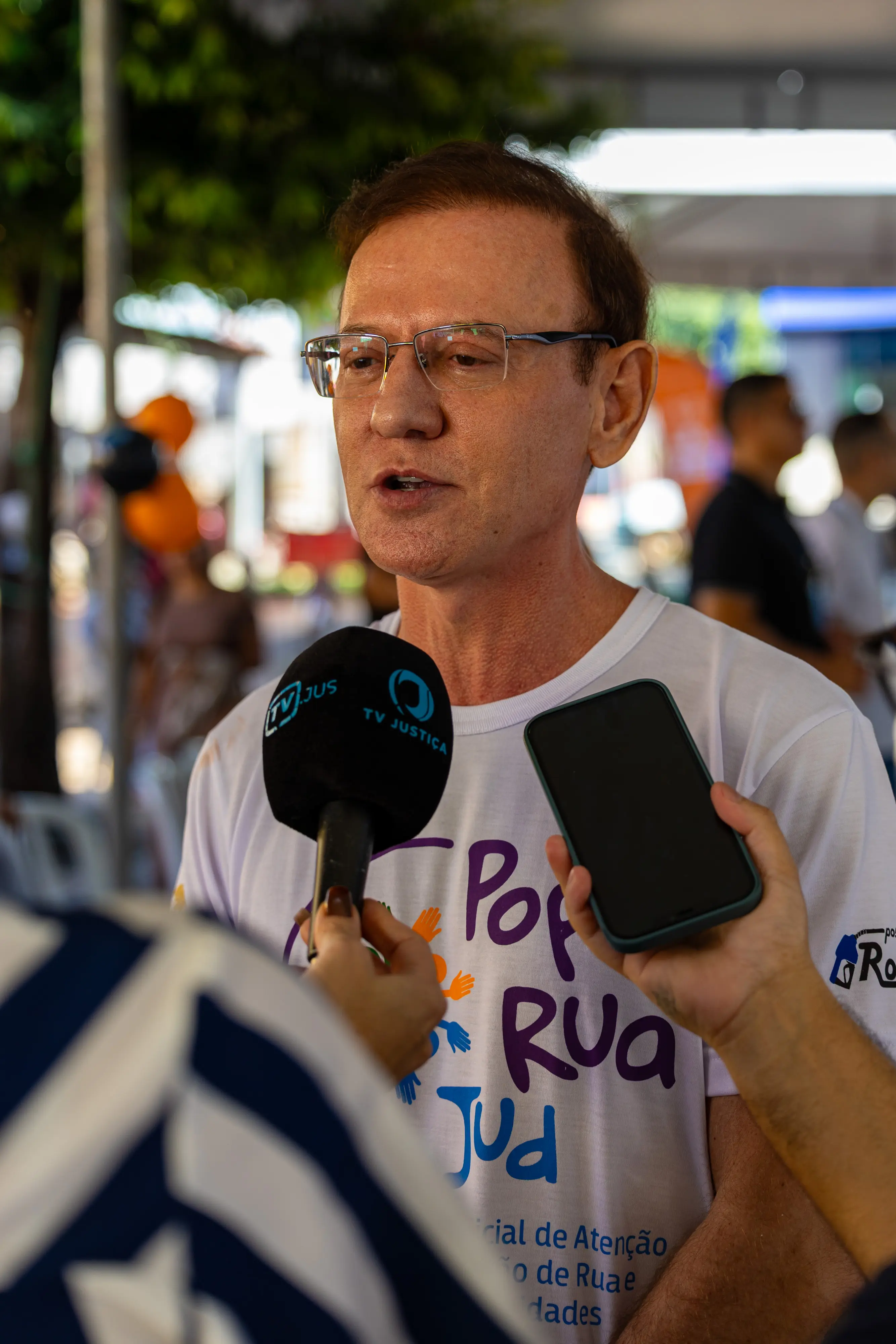 Homem de óculos e camiseta branca com logo "Pop Rua Jud" dá entrevista. Um microfone da TV Justiça e um celular estão posicionados à frente dele para a gravação.