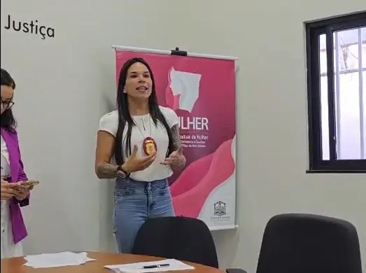 Mulher branca de cabelos longos escuros, com calça jeans e blusa branca com distintivo no pescoço, falando e gesticulando. 