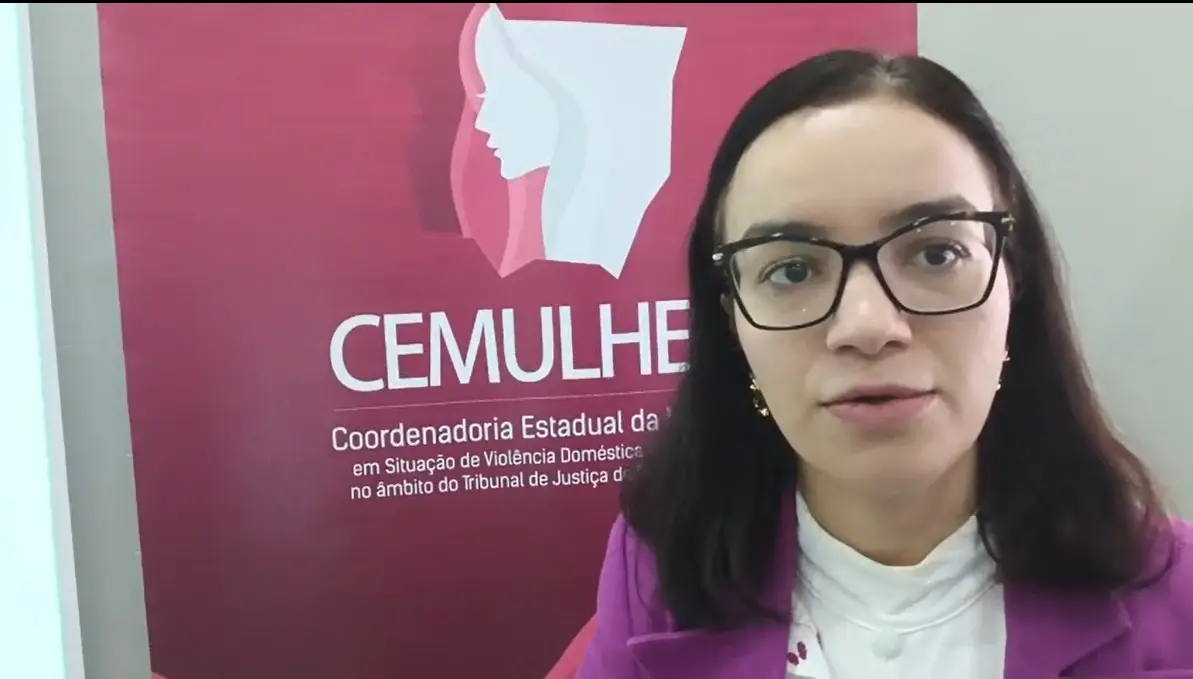 Mulher branca, de cabelos médios, com óculos e blazer roxo falando diante do banner da CEMULHER. 