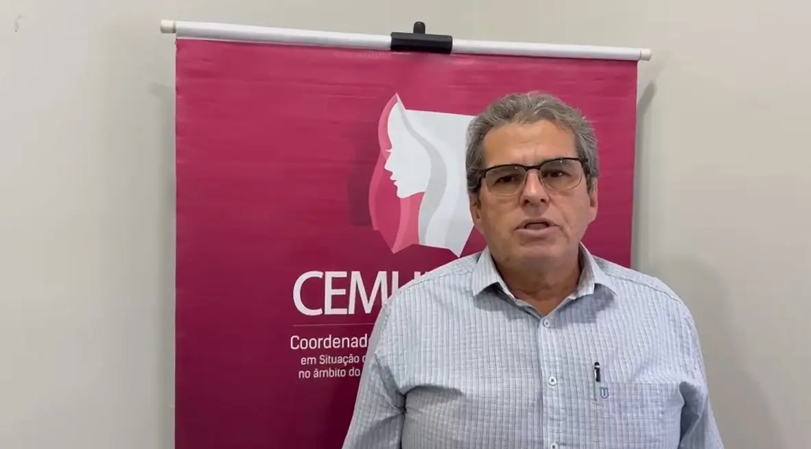 Homem branco, de cabelos grisalhos, com óculos e camisa xadrez clara, falando diante de um banner da CEMULHER.
