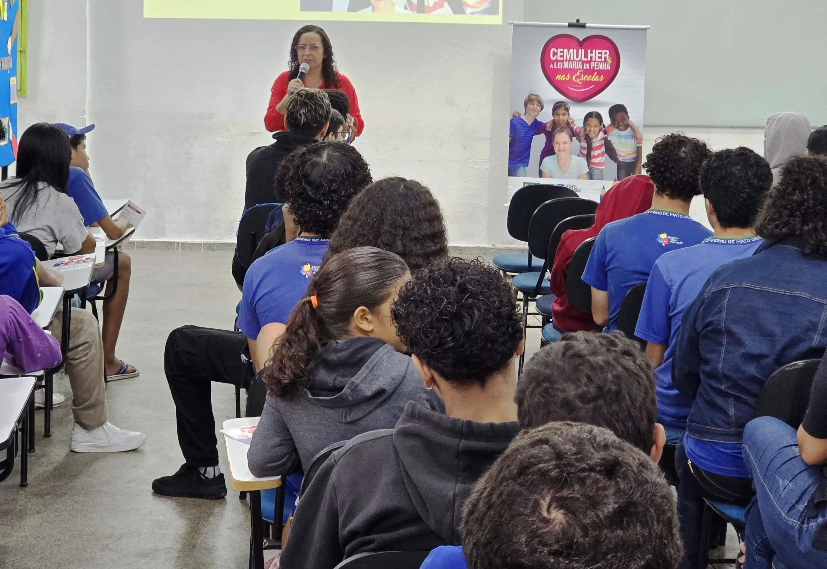 A assistente social da Cemulher está em pé, dentro de uma sala de aula, falando com estudantes, que estão de frente para ela, sentados e cadeiras