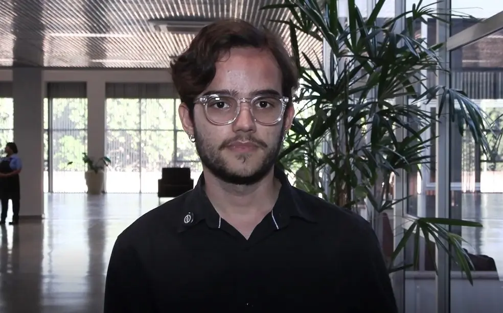 A imagem mostra o acadêmico Sebastião. Ele é um jovem de pele clara, olhos, cabelos e barba escuros. Ele usa óculos de grau e veste camisa preta. Ele está num dos corredores amplos do Complexo