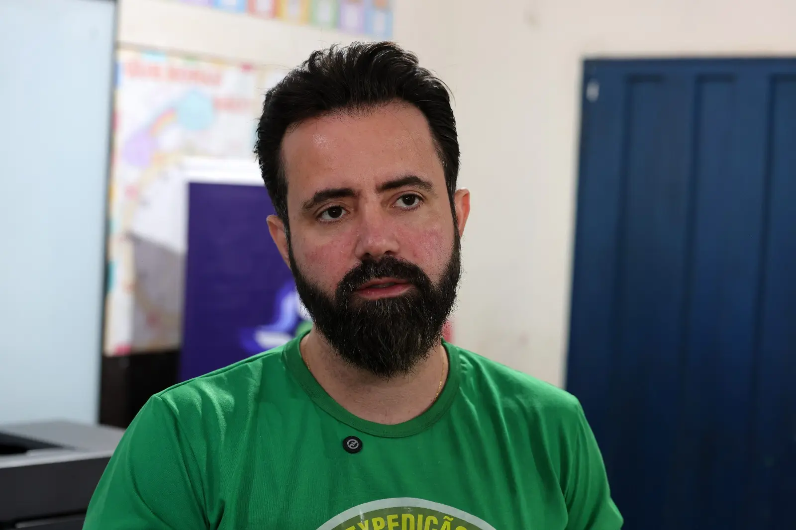 Juiz federal Flávio Fraga da Silva é um homem de pele clara e barba escura aparada olha ligeiramente para o lado. Ele veste uma camiseta verde-bandeira de gola careca e o fundo mostra um ambiente interno com portas e cartazes.