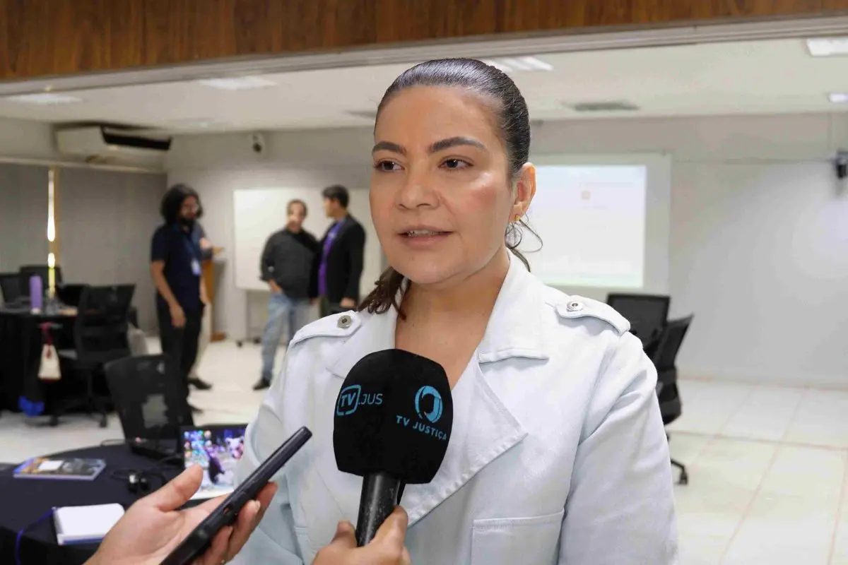 Foto que mostra a servidora Hellen Graciosa, uma mulher branca, de cabelos presos, vestindo um blazer claro, falando ao microfone da TV JUS em uma sala de eventos.