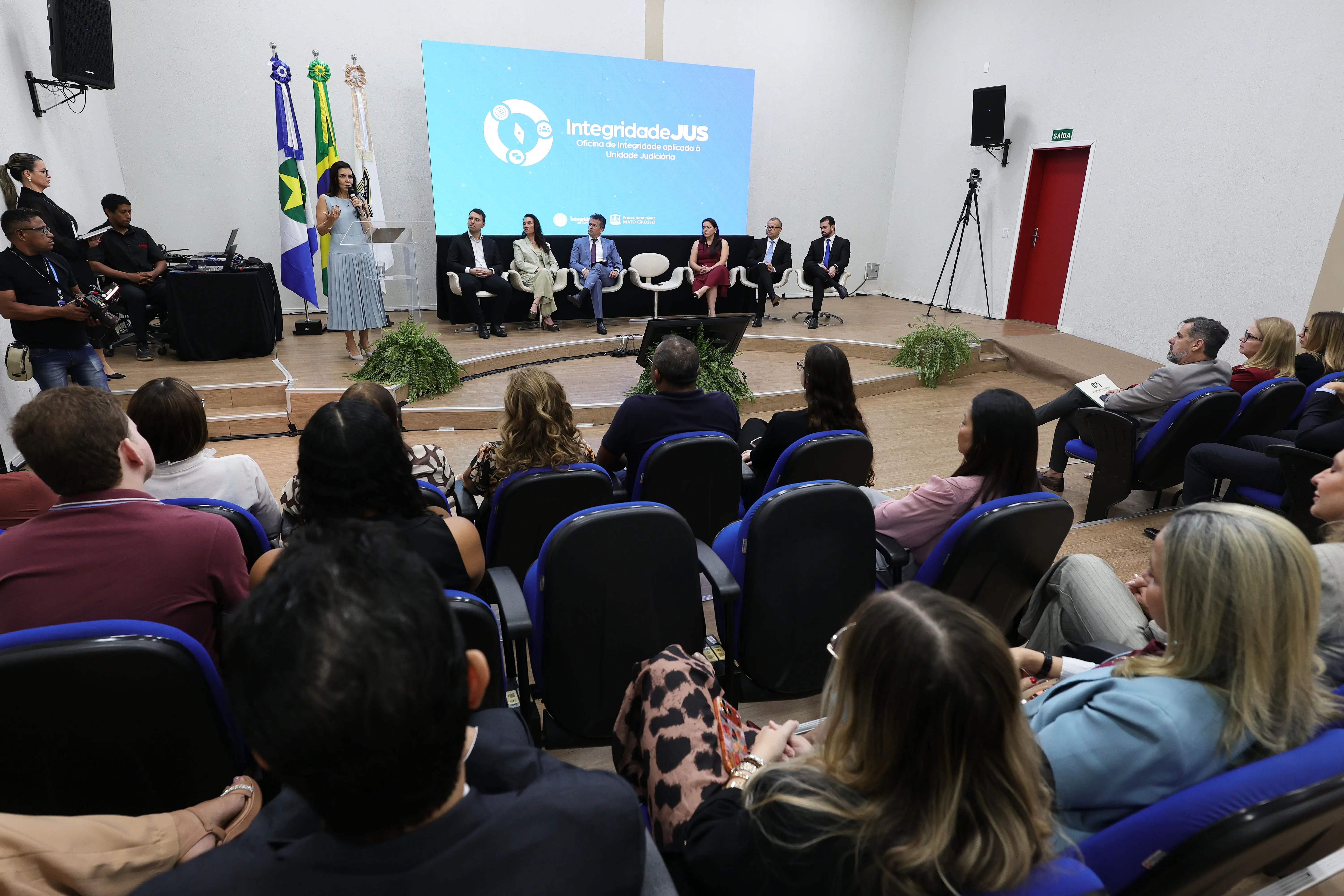 Vista geral do auditório lotado. Em primeiro plano, a plateia de costas em poltronas azuis. Ao fundo, o palco com uma palestrante no púlpito, composição da mesa e telão "IntegridadeJUS".