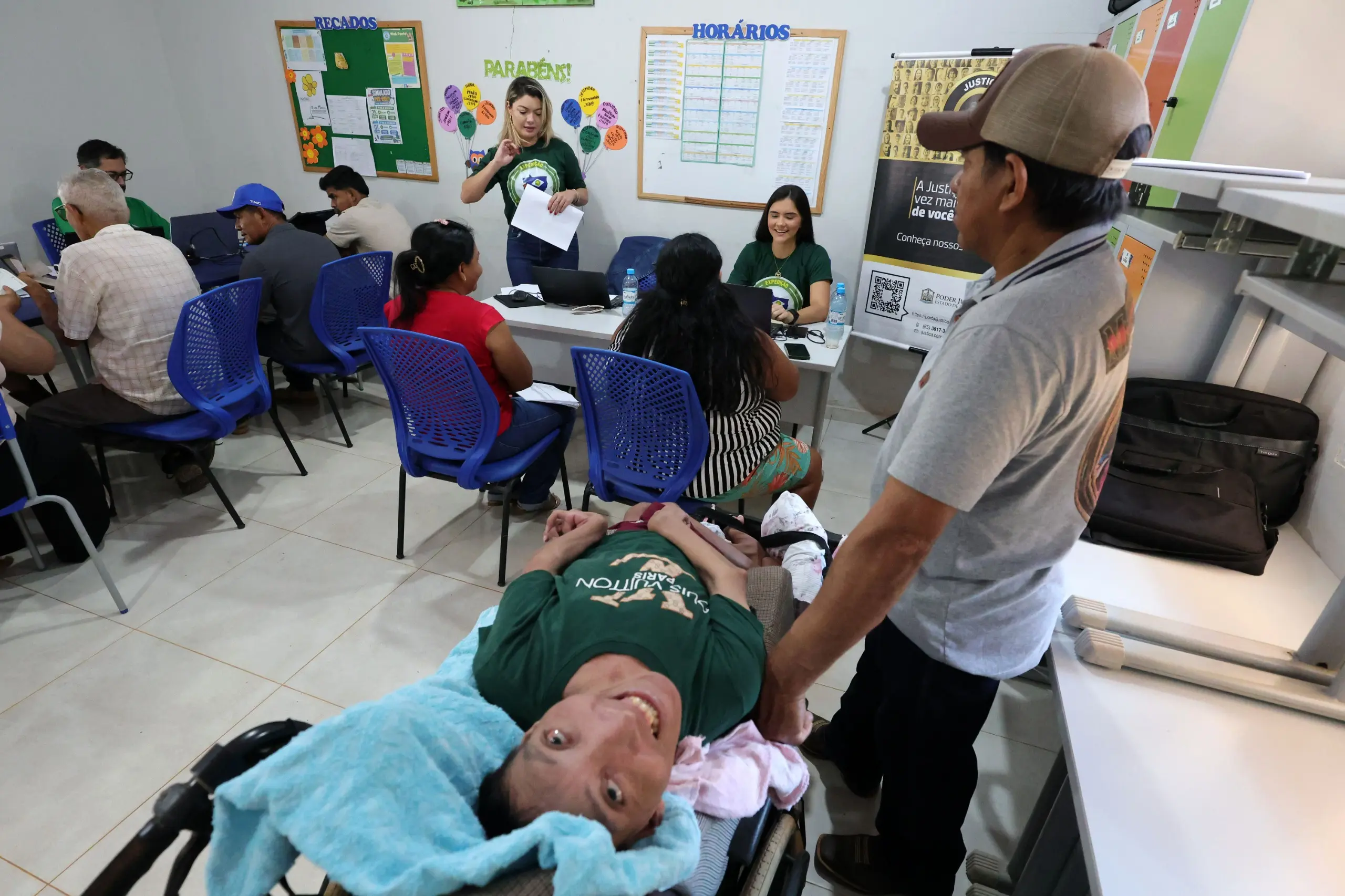 Pessoa em cadeira de rodas é conduzida por um homem dentro de uma sala de atendimento. No local, servidores atendem outras pessoas sentadas em mesas com computadores.