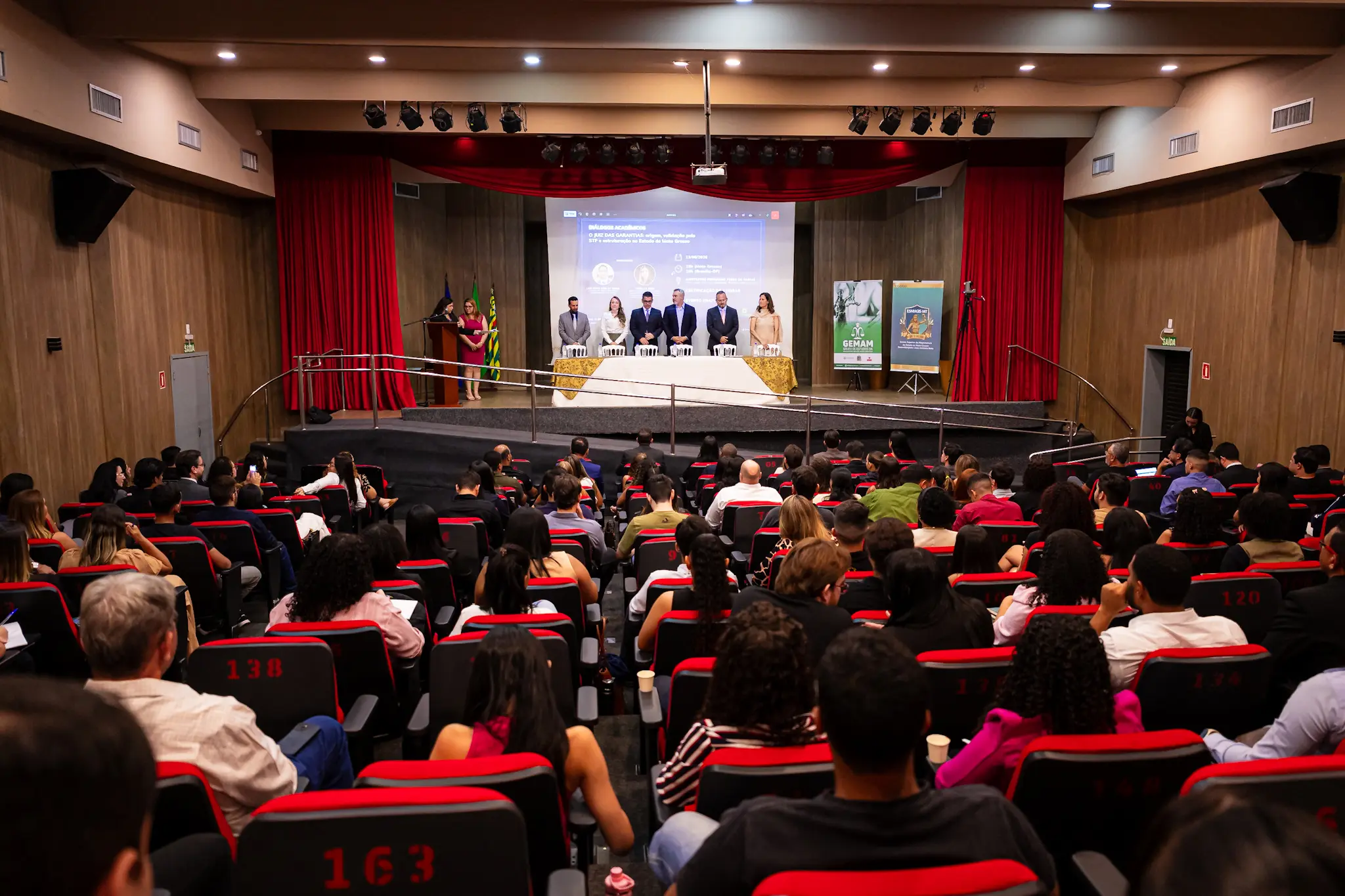 Em vista ampla, auditório lotado com assentos vermelhos. No palco, pessoas de traje formal estão de pé atrás da mesa principal sob luzes fortes.
