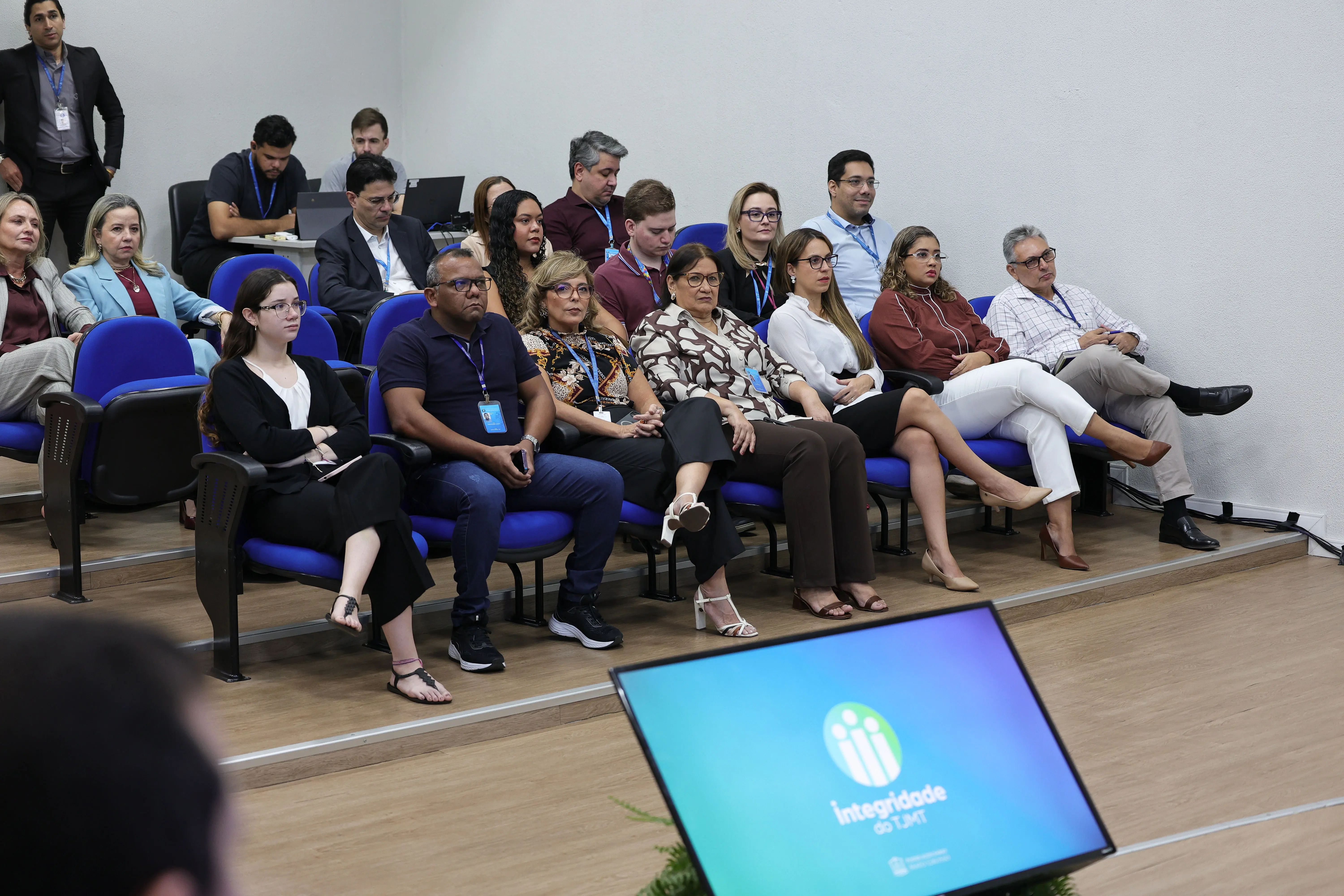Pessoas sentadas em poltronas azuis de um auditório acompanham a apresentação. Em primeiro plano, um monitor exibe o logotipo "Integridade do TJMT" em fundo azul.