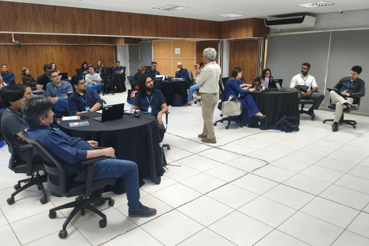 Foto horizontal com a visão geral de uma sala de treinamento, com pessoas sentadas em mesas redondas pretas. Um homem em pé, no centro, fala ao grupo.