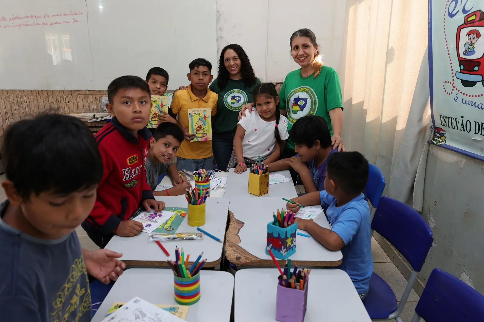 Crianças reunidas em mesas escolares com potes de lápis de cor e desenhos. Ao fundo, duas mulheres com camisetas verdes da expedição sorriem em pé ao lado de alguns alunos que mostram livretos.