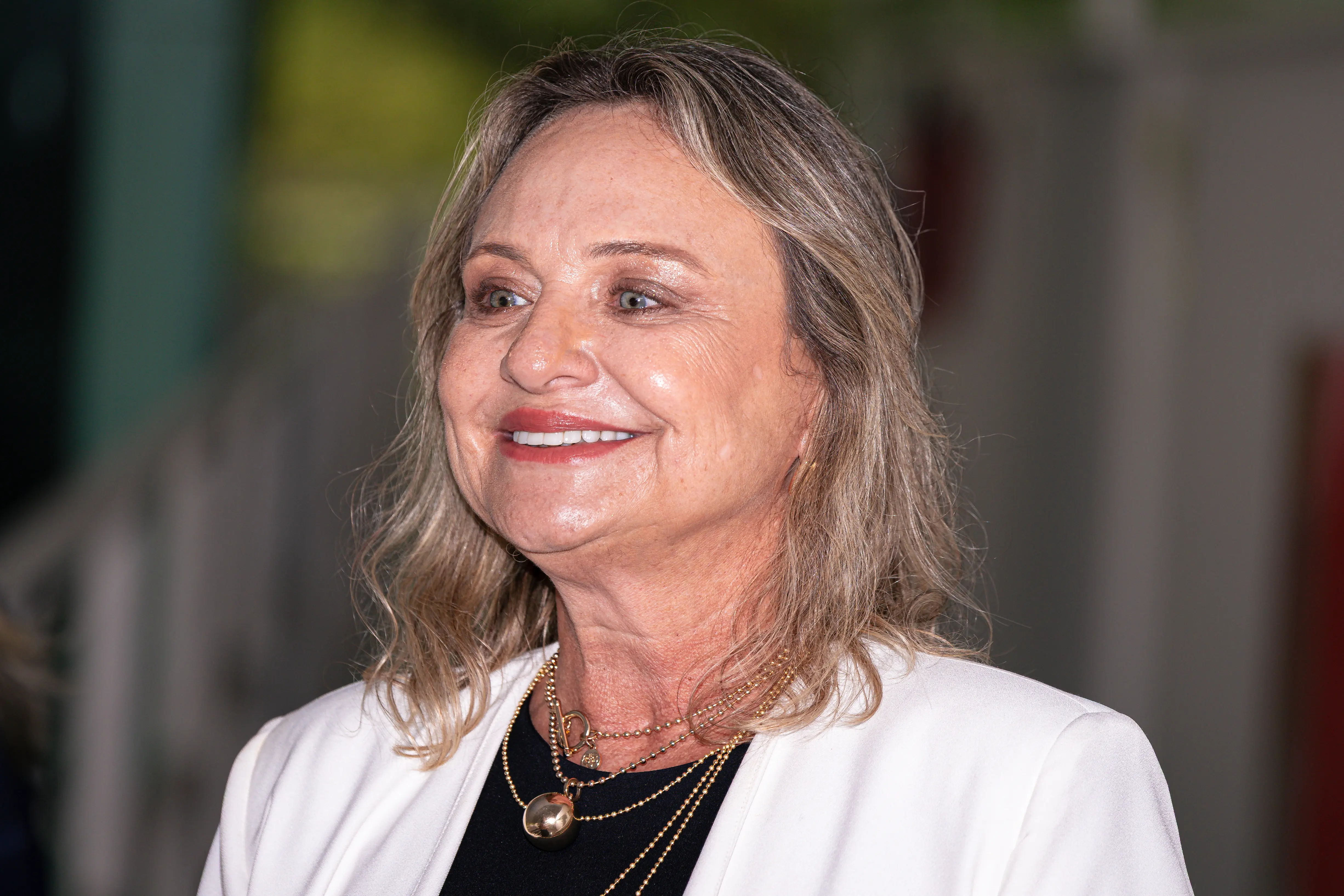 Mulher de pele clara sorrindo, com cabelos loiros ondulados na altura dos ombros. Usa blazer branco sobre blusa preta e colares dourados com pingente esférico. Olhar direcionado para a esquerda.