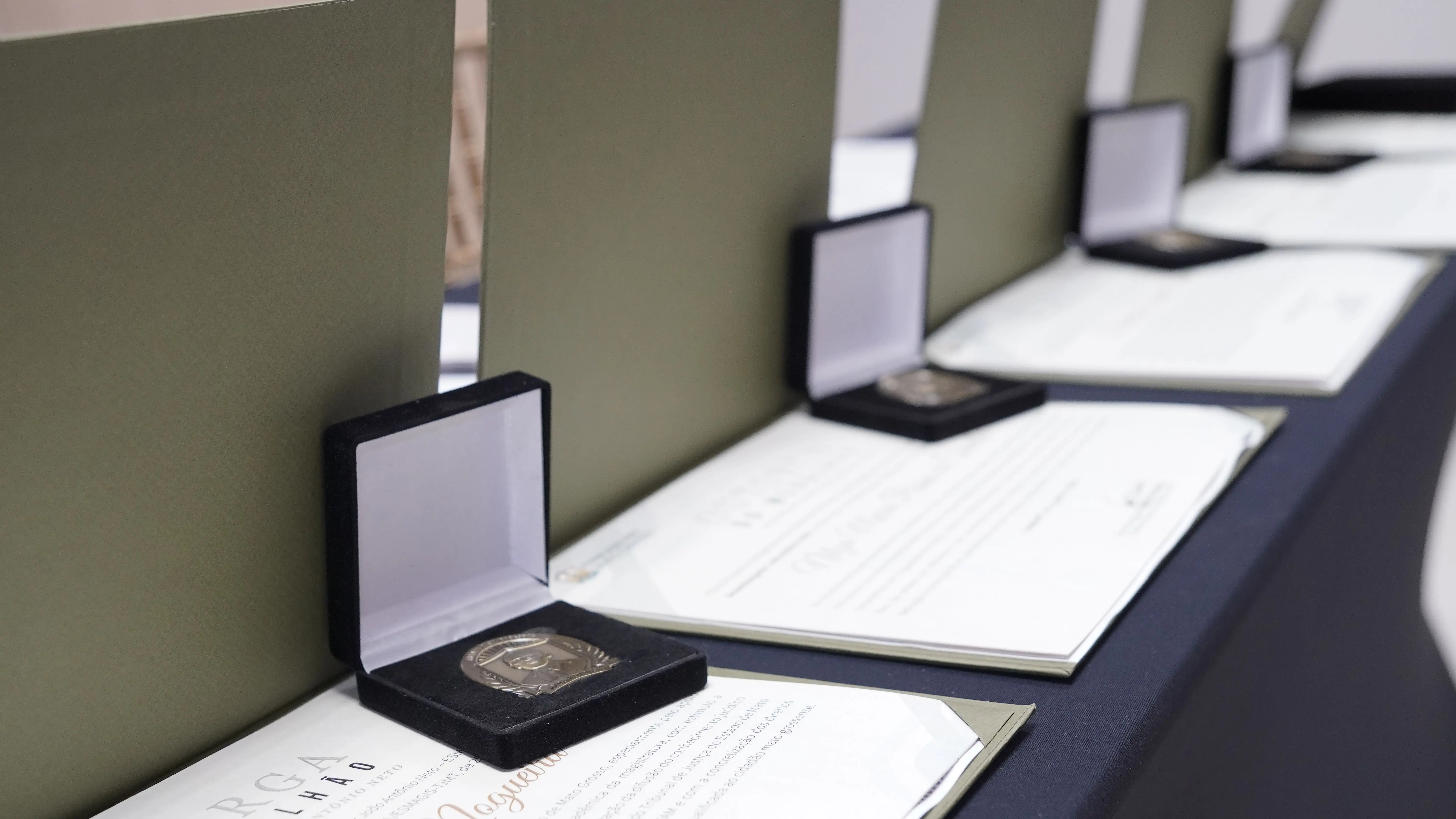 Imagem em close mostra uma fileira de caixas abertas contendo medalhas, dispostas sobre uma mesa ao lado de certificados impressos. As caixas são escuras por fora e claras por dentro, alinhadas lado a lado em perspectiva. Os certificados estão parcialmente visíveis sob as caixas.