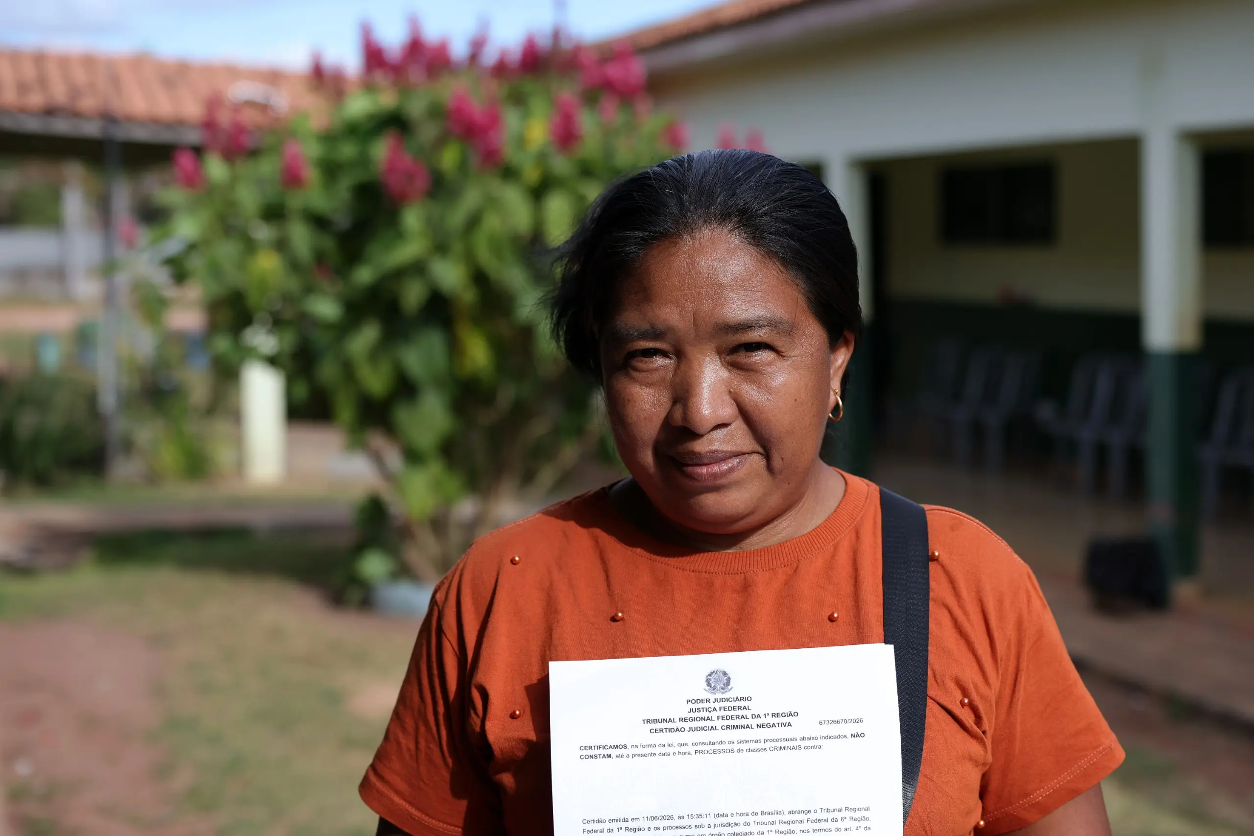 Foto horizontal que mostra a boliviana Gerônima Chube sorrindo timidamente para a foto e mostrando um documento. Ela está no pátio de uma escola. Ela é uma mulher com traços bolivianos, pele marrom, cabelo preto, liso e preso para trás, usando camiseta marrom.