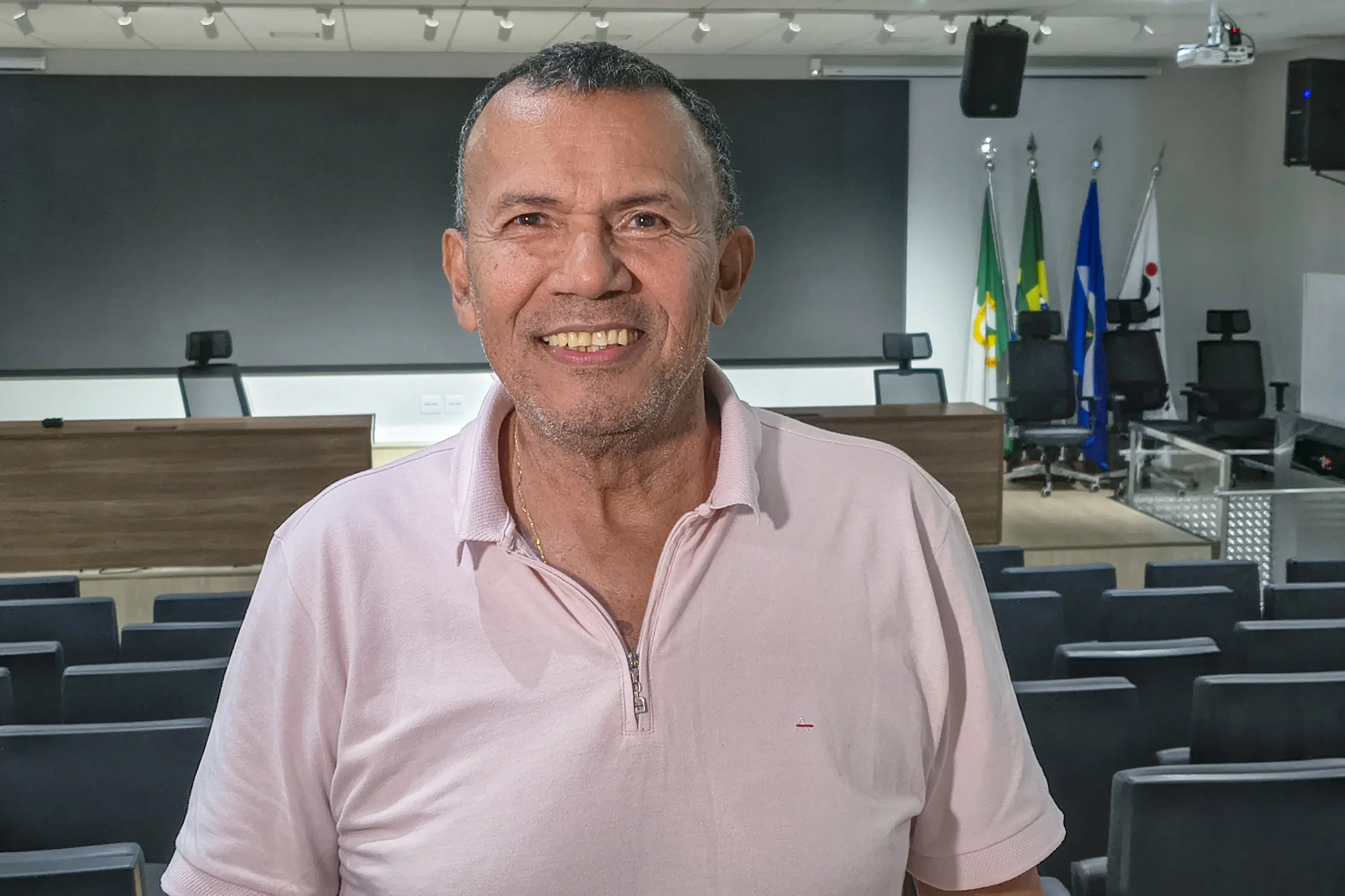 Homem de cabelos grisalhos sorri, vestindo polo rosa claro. Está em auditório vazio, com cadeiras ao fundo, bandeiras e um púlpito de madeira ao lado direito da imagem.