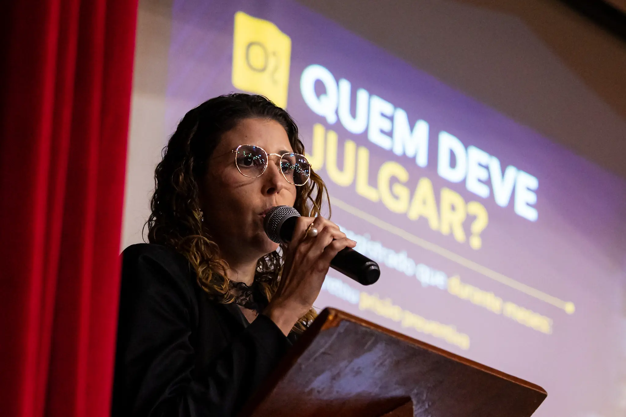 Uma mulher de cabelos ondulados e óculos fala ao microfone no púlpito, vista de perfil. Ela veste roupa escura e segura o microfone próximo à boca