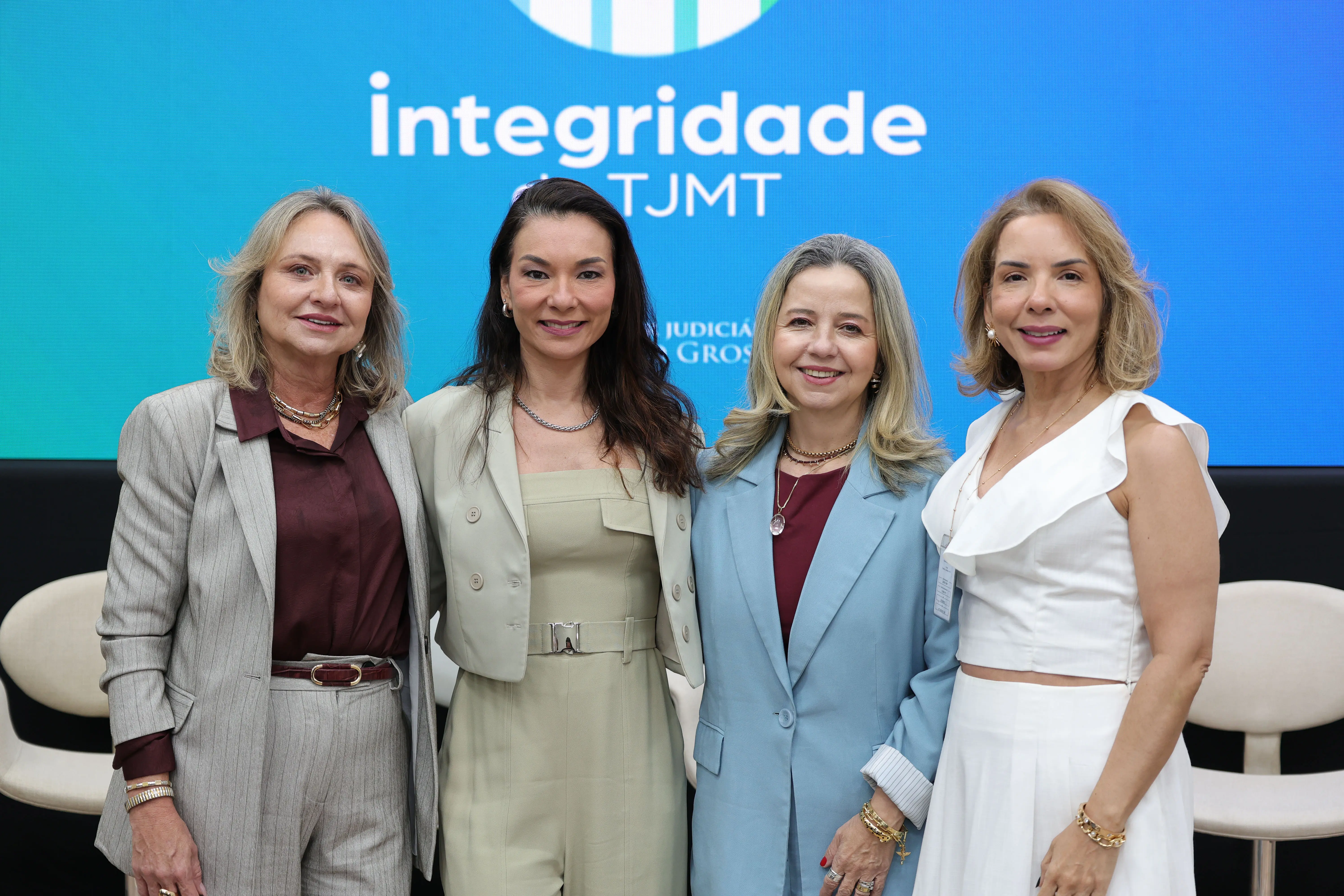 Quatro mulheres sorriem lado a lado em frente a um telão azul com a palavra "integridade do TJMT". Elas vestem trajes formais em tons de cinza, bege, azul-claro e branco.