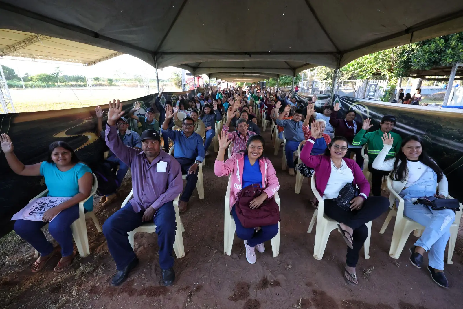 Dezenas de pessoas estão sentadas em cadeiras plásticas sob uma grande tenda de lona. Elas olham para a câmera e acenam com uma das mãos levantadas. O chão é de terra batida.