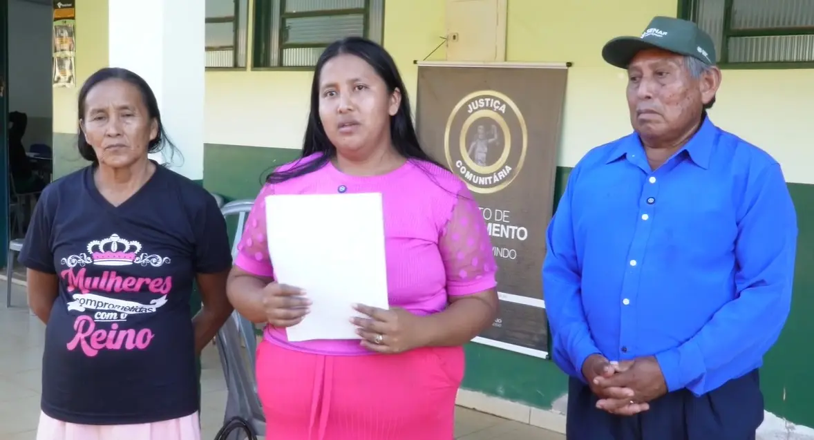 Três pessoas indígenas em pé: à esquerda, uma mulher de blusa preta; ao centro, uma mulher de rosa segurando papéis; à direita, um homem de camisa azul e boné. Ao fundo, um banner 