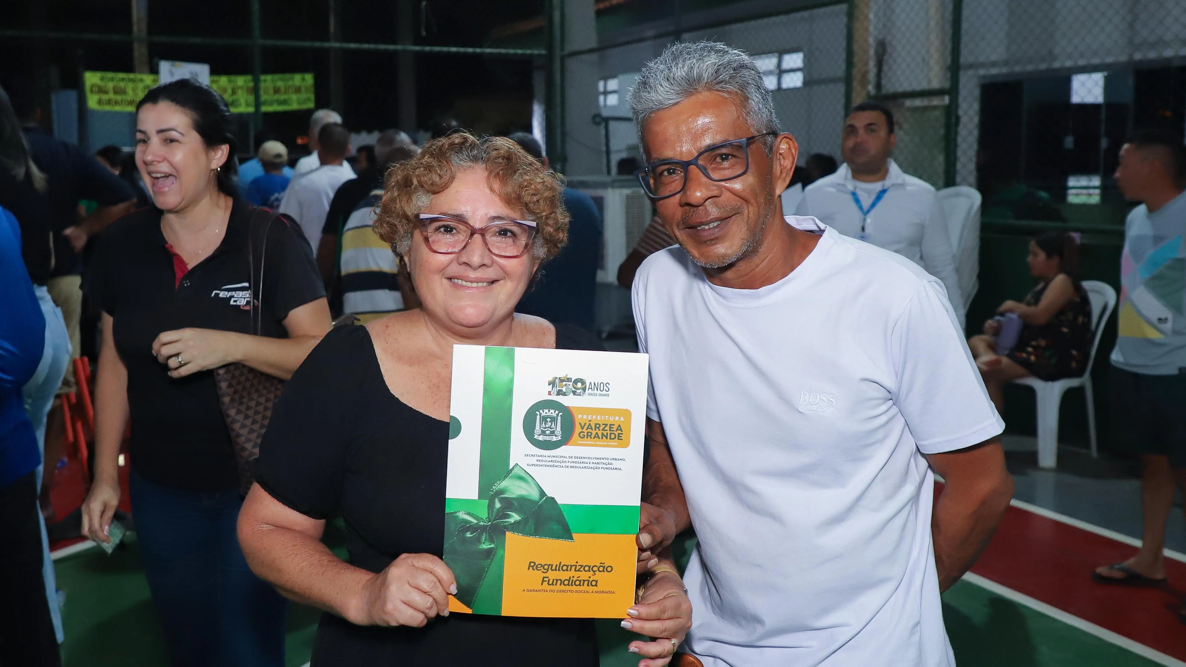 Moradores do bairro Jardim Manaíra exibem o documento de regularização fundiária durante cerimônia realizada na Escola Municipal de Educação Básica (EMEB) Ednilson Francisco Kolling, em Várzea Grande.