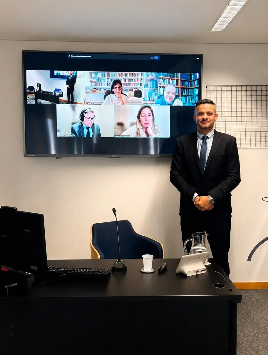 Um homem de terno preto e gravata azul posa em pé à direita de uma mesa de escritório preta. No centro da parede branca ao fundo, uma grande TV exibe uma videoconferência com quatro outras pessoas em janelas separadas.