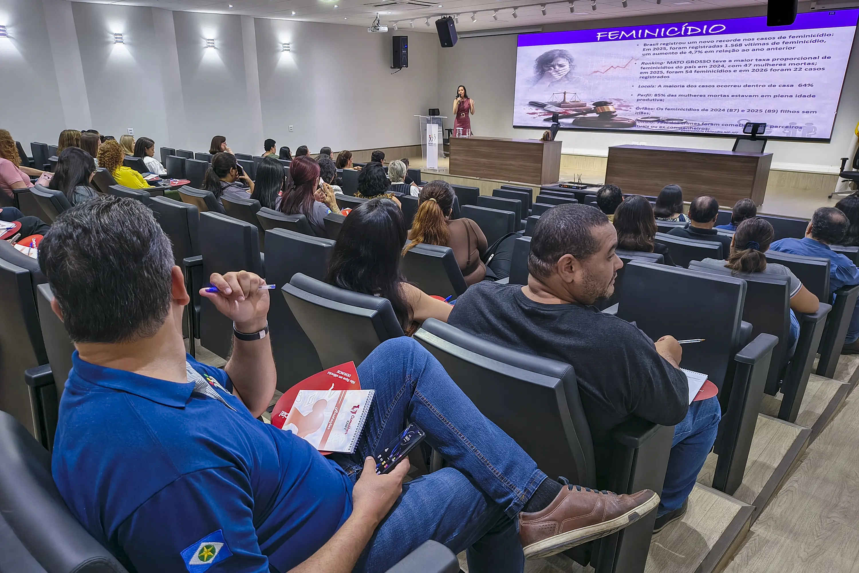 Plateia acompanha palestra sentada, de costas para a câmera. Ao fundo, telão exibe dados sobre feminicídio em Mato Grosso e palestrante fala ao microfone no púlpito.