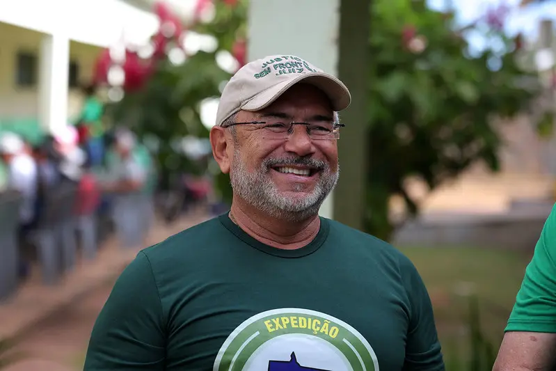 Homem sorridente de óculos, barba grisalha, boné bege e camiseta verde escrito 