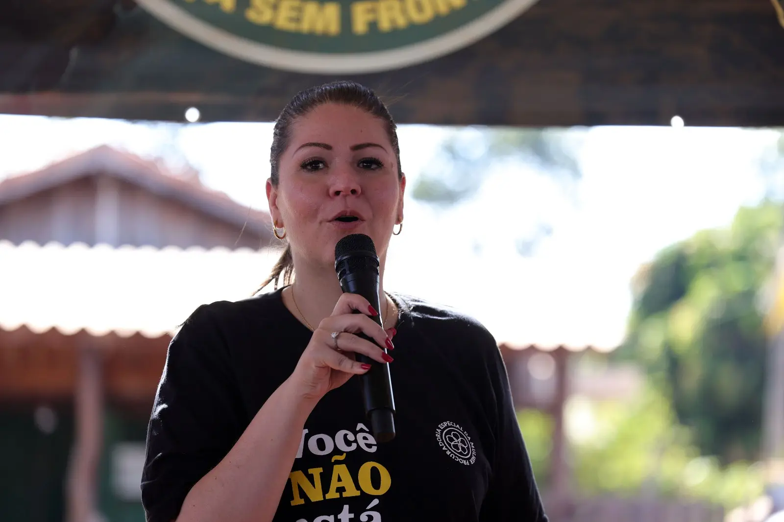 Mulher de pele clara fala ao microfone que segura com a mão direita. Ela usa cabelos presos, brincos dourados e camiseta preta com letras amarelas e brancas, além de um logotipo circular.